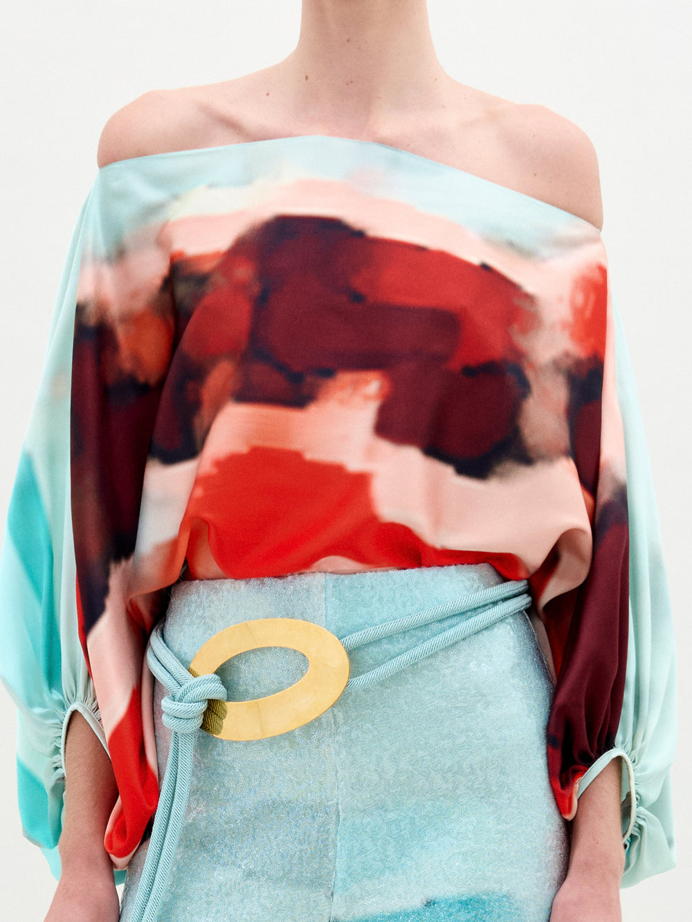 A woman wears the Bellagio Blouse, an off-shoulder red and aqua abstract print silk top with an oversized fit, paired with light blue pants featuring a large circular yellow belt buckle, against a plain background.