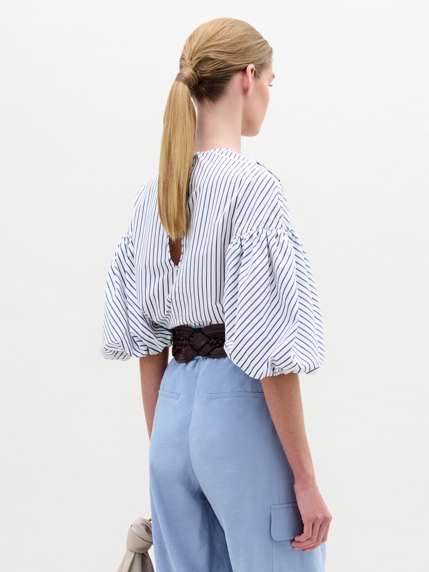 A woman wearing the Belle Blouse, featuring blue floral embroidery and voluminous sleeves, paired with light blue high-waisted pants, a wide brown belt with a large circular buckle, and holding a small handbag.