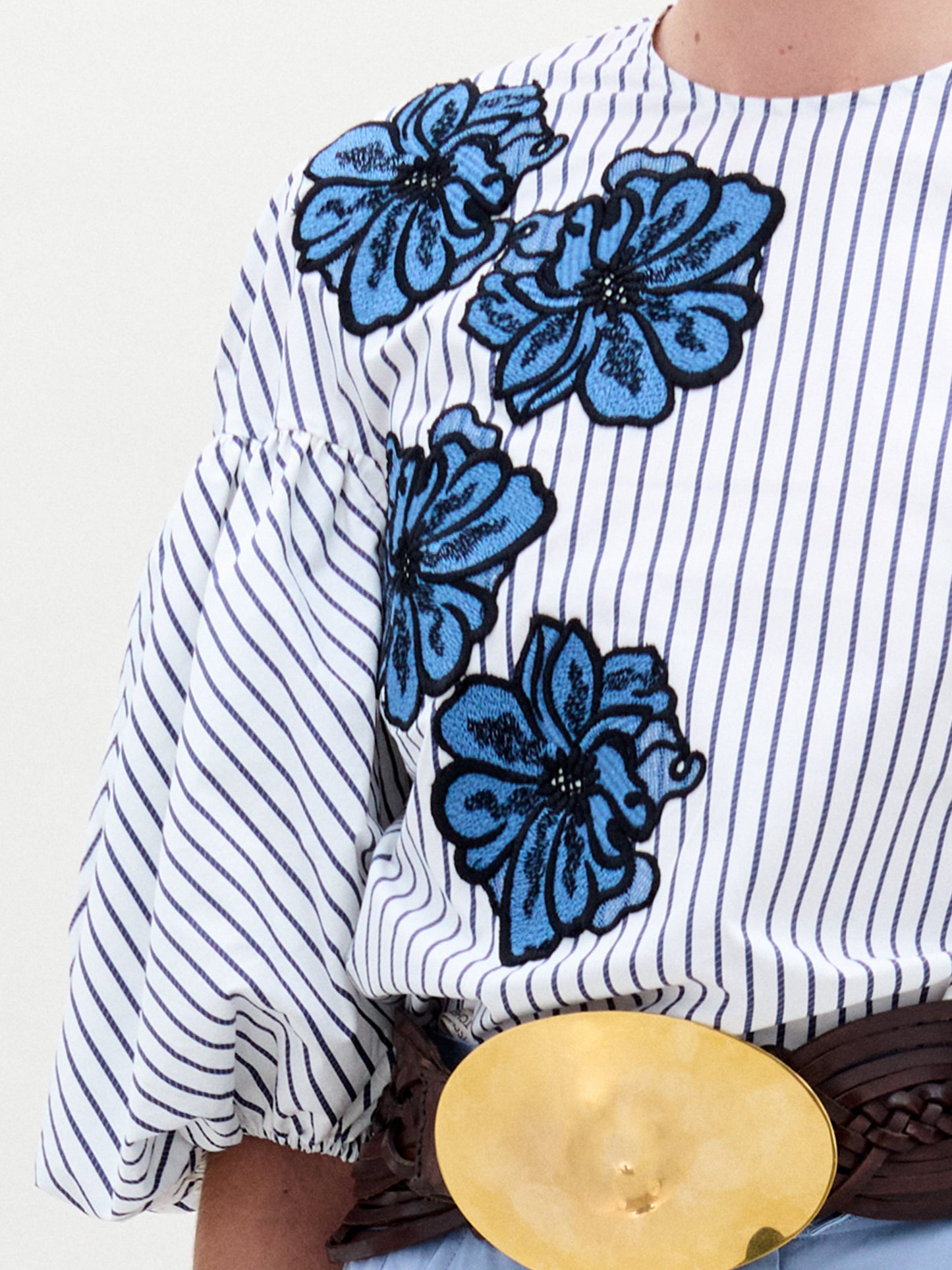 A woman wearing the Belle Blouse, featuring blue floral embroidery and voluminous sleeves, paired with light blue high-waisted pants, a wide brown belt with a large circular buckle, and holding a small handbag.