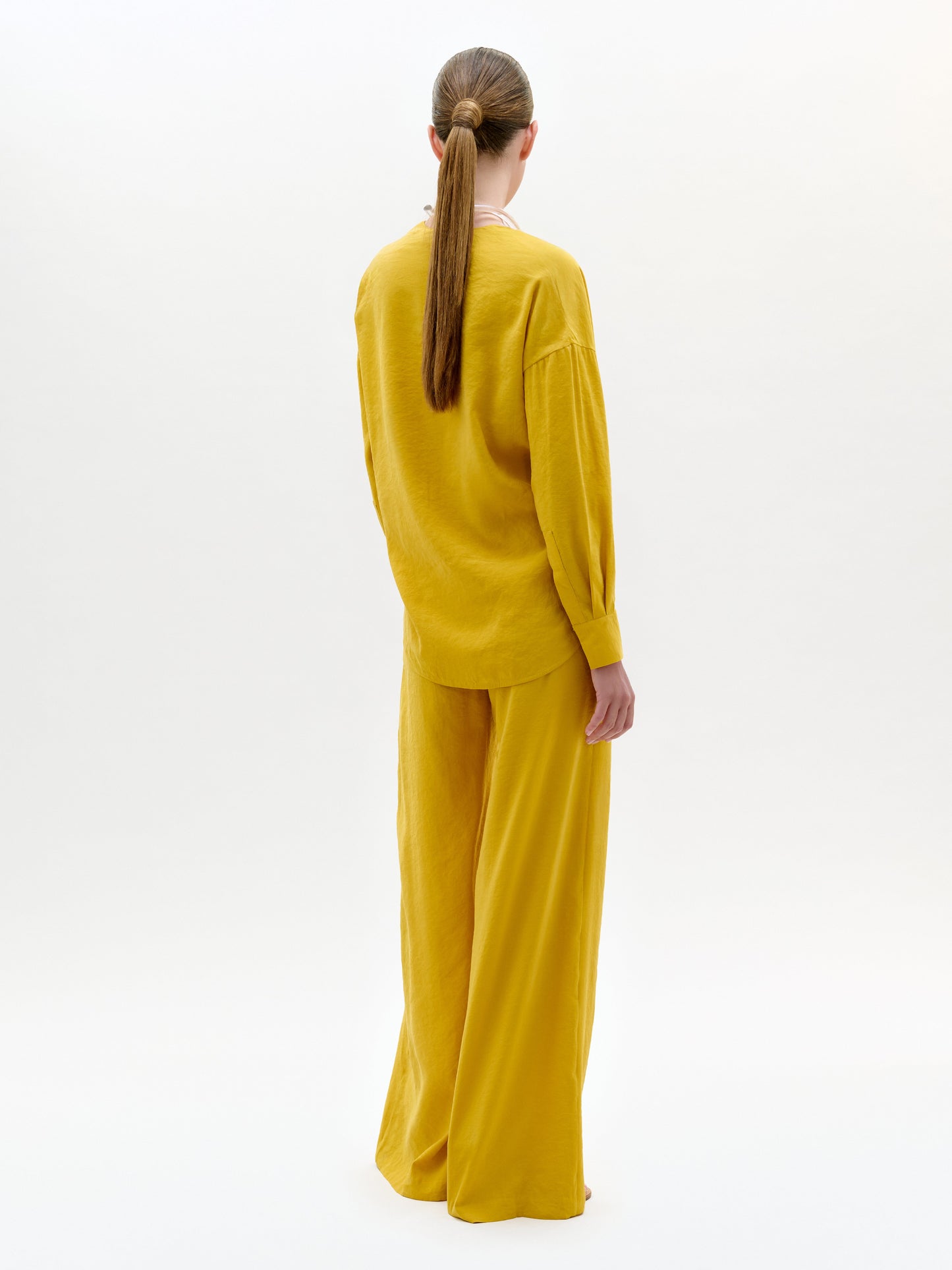 A women stands confidently in a mustard yellow jumpsuit with a deep V-neck and cinched waist, against a plain white background, exuding elegance.
