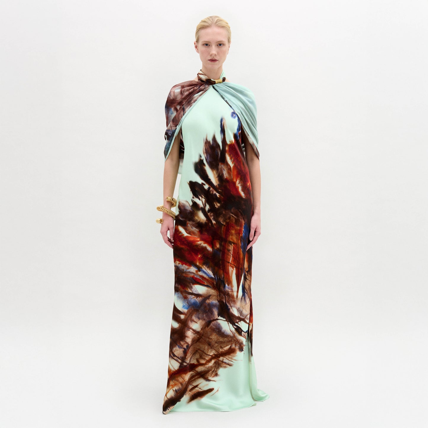 A woman stands against a white background wearing the Carlina Dress, a floor-length light blue maxi made from Italian fabric with a brown and red feather-like pattern and a matching draped shoulder cape.