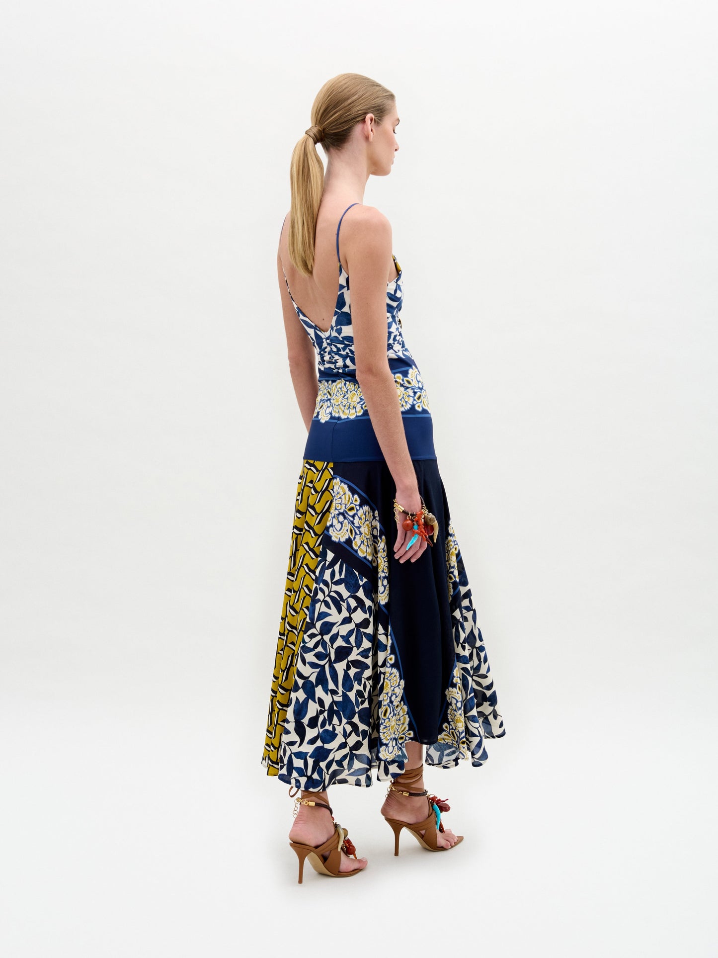 A woman poses against a white background wearing the Casiana Dress, a sleeveless blue, yellow, and white patterned midi with a flared jersey skirt, styled with brown strappy sandals.