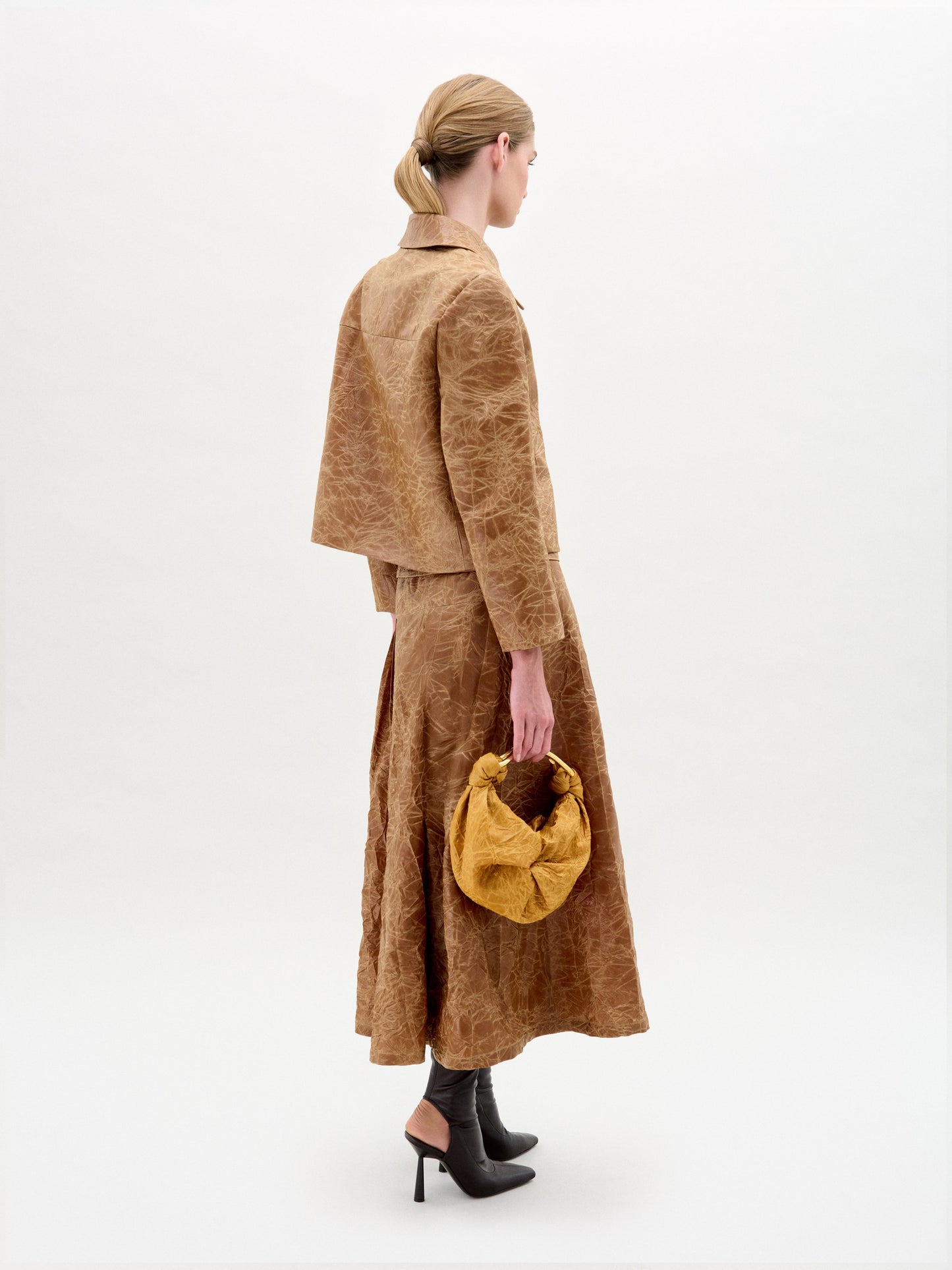 A woman wears the Olga Skirt in brown with a matching textured A-line jacket, gold accent belt, dark ankle boots, and a minimal background.