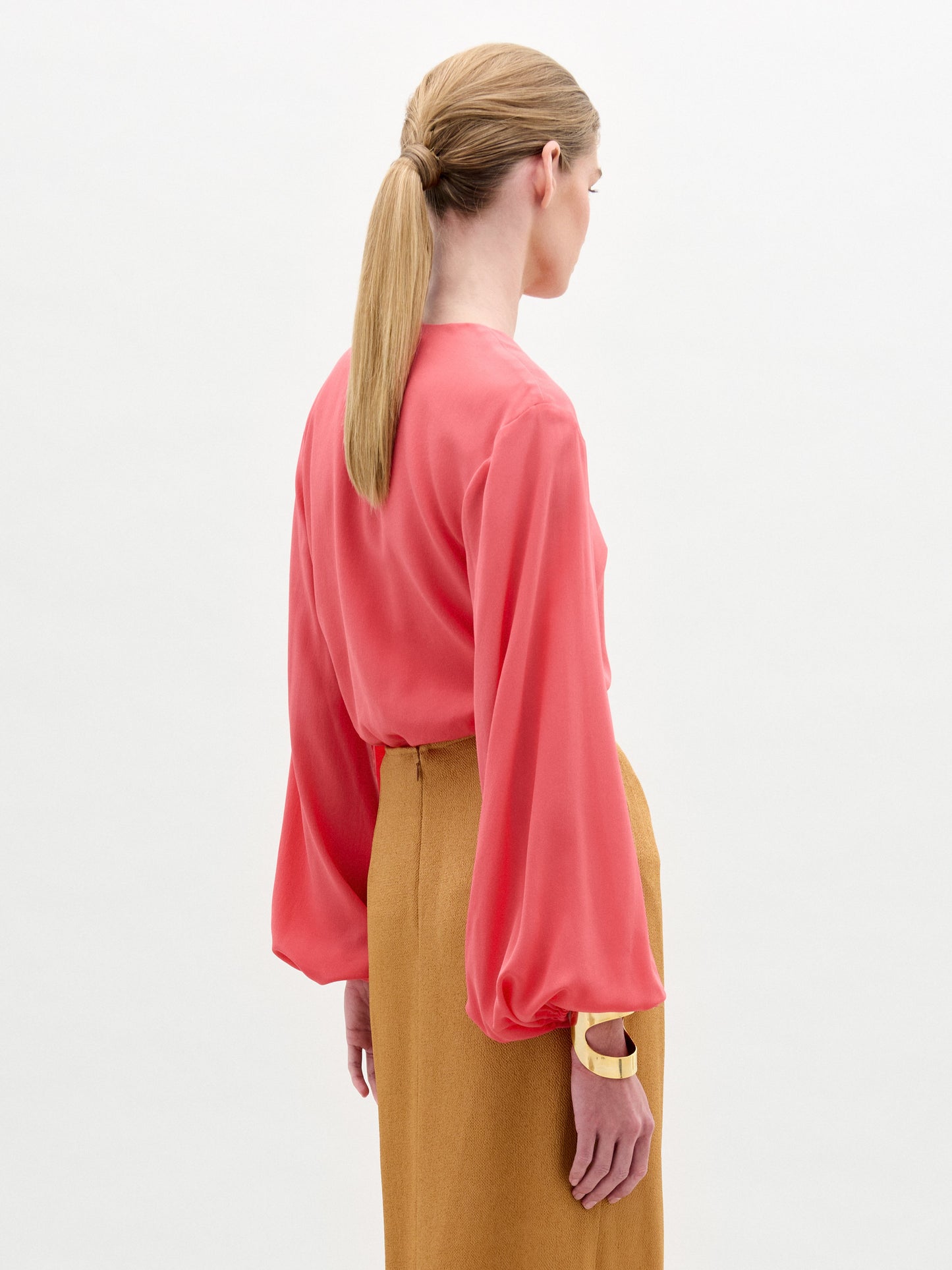 A woman faces away, wearing the Covilha Blouse—a coral pink silk top with billowy sleeves and a keyhole neckline—paired with a high-waisted camel skirt. Her hair is in a low ponytail against a plain white background.