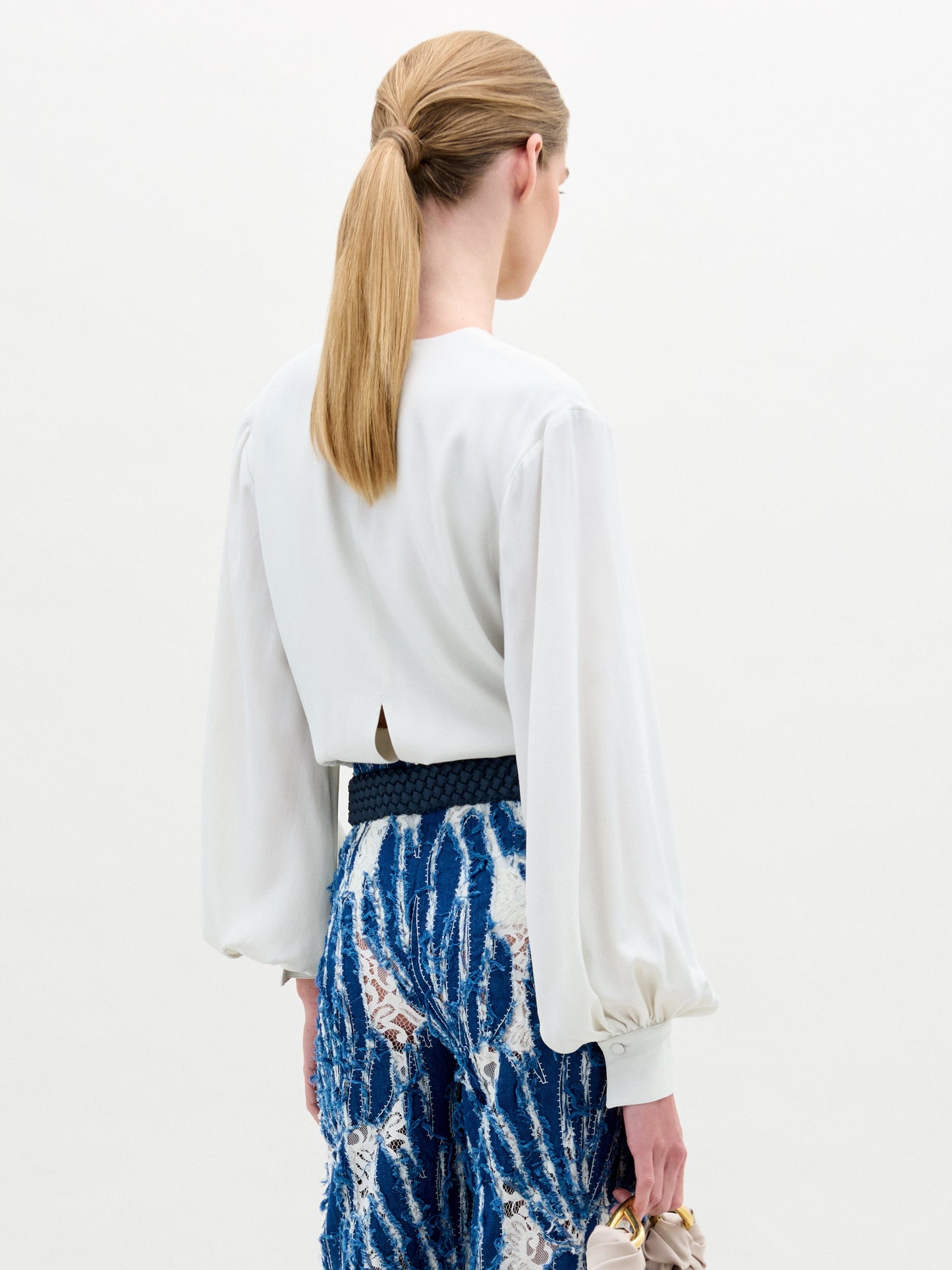 A woman poses against a plain background wearing the Covilha Blouse, featuring a keyhole neckline and balloon sleeves, styled with high-waisted blue patterned pants and holding a small beige bag.