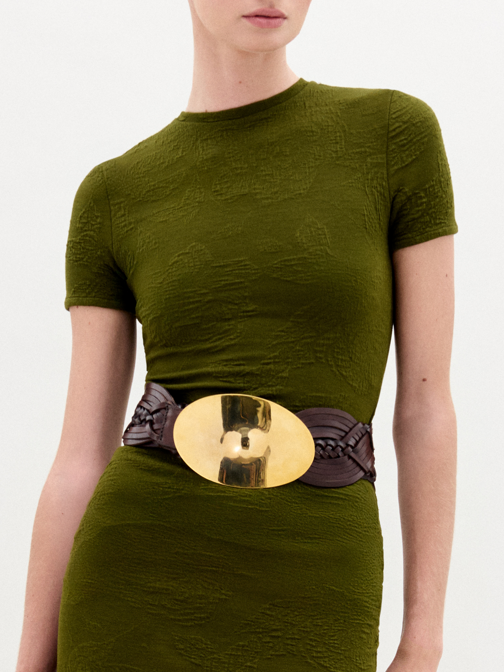 The Clarissa Belt, featuring brown braided leather and a large shiny gold buckle, is displayed coiled on a plain white background.