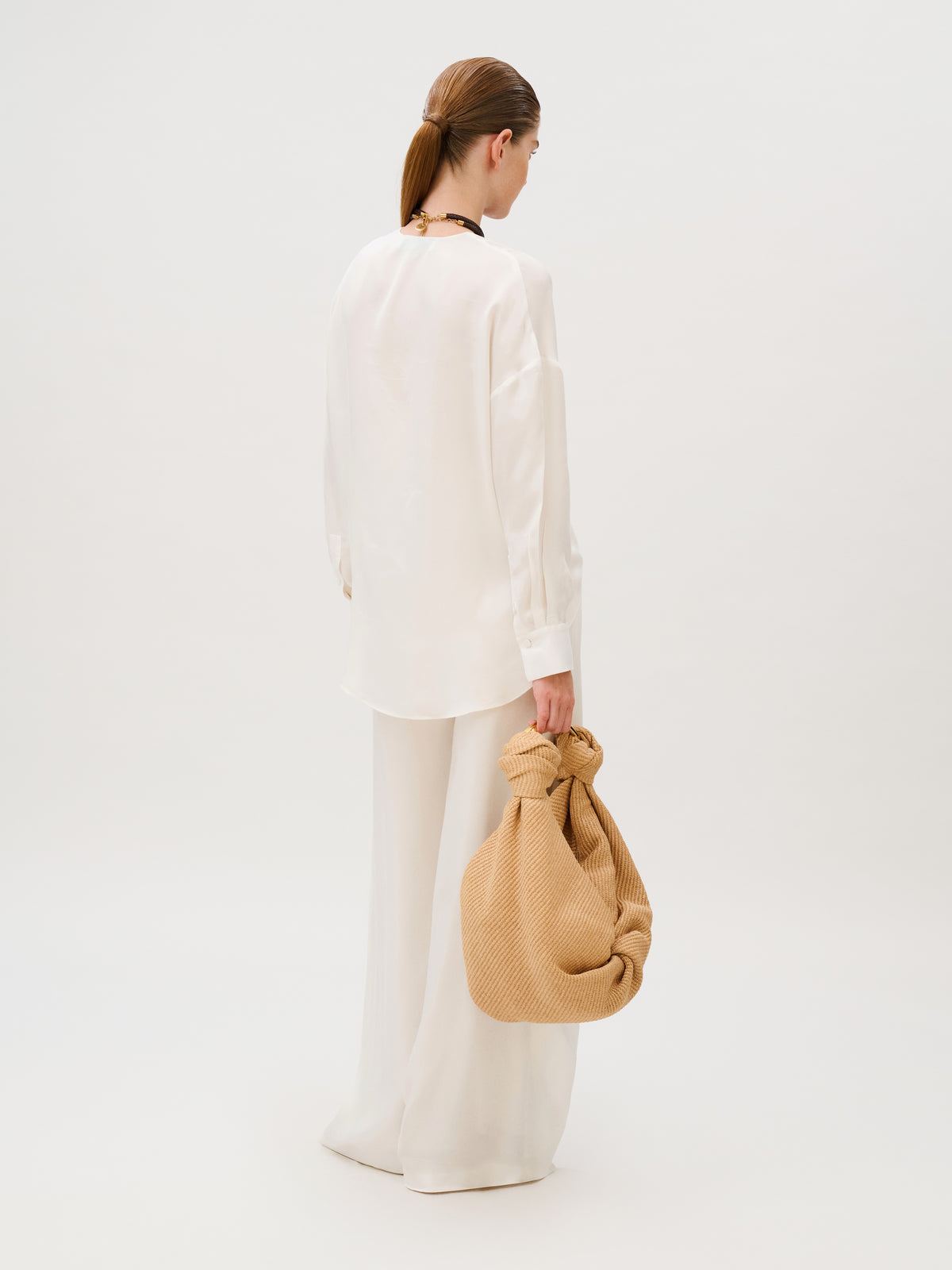A person seen from the back wearing a long-sleeved white shirt and matching white Canturipe Pant, holding a large, knotted tan fabric bag.