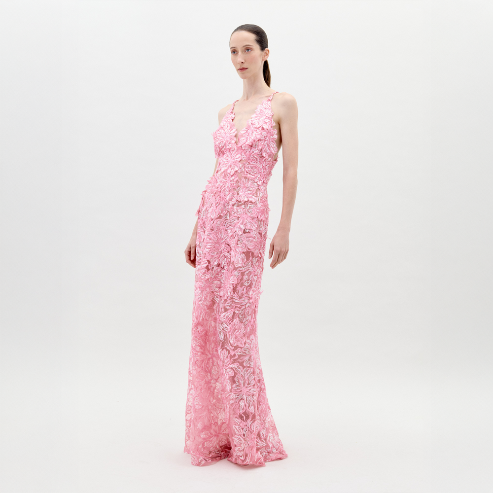 A person stands wearing the Daira Dress Pink, a long dress with thin straps and a beautiful floral pattern. The background is plain and light-colored. This elegant dress is now available for pre-order and will ship by February 1st, 2025.