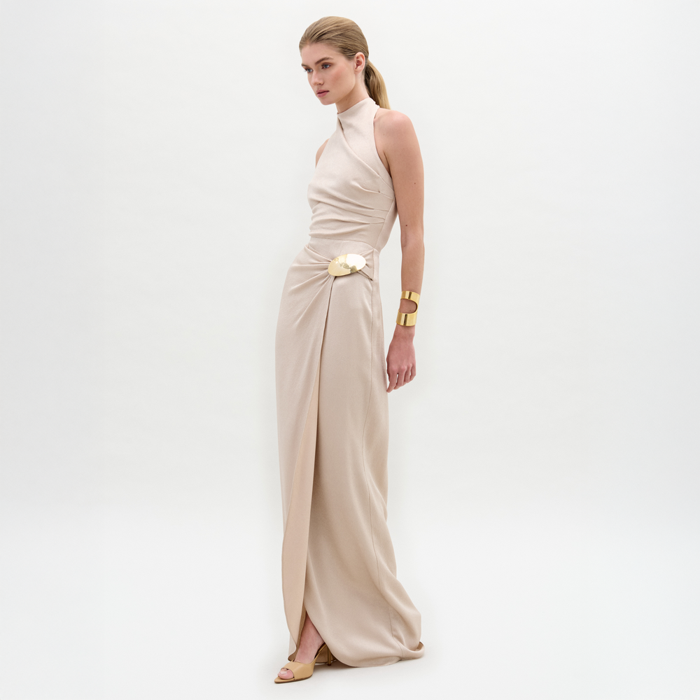 A woman wears the Davinia Dress, a cream, floor-length halter gown with a floral accent at the waist, paired with a gold cuff bracelet and beige shoes, standing against a plain white background.