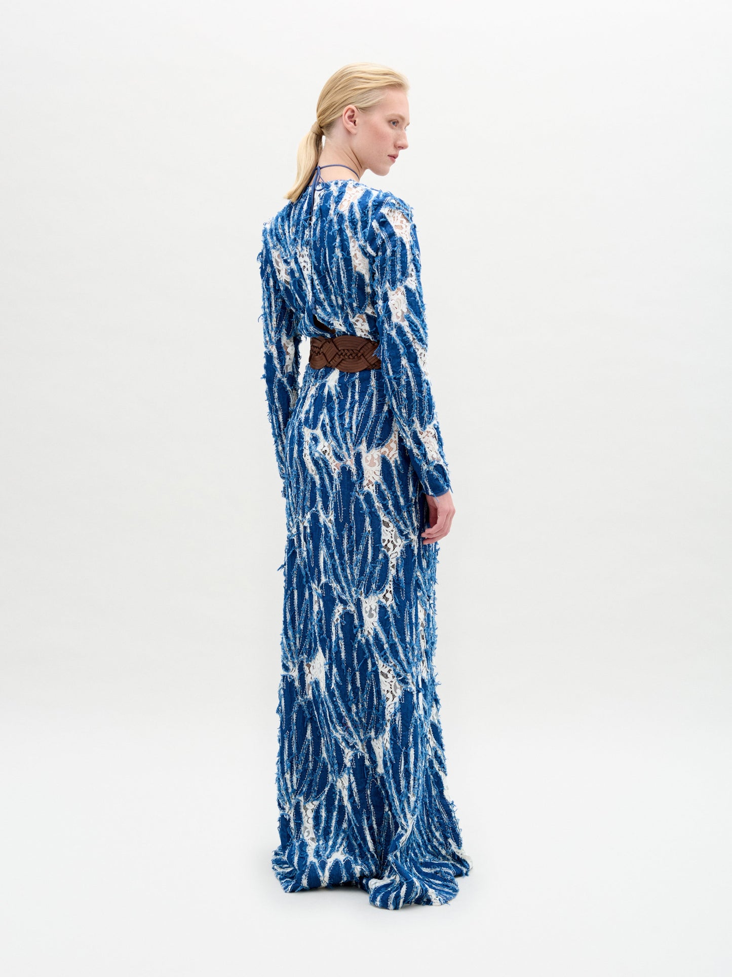 A person stands against a plain background wearing the Ebba Dress, a long blue and white patterned dress with a brown belt, high neckline, and subtle criss-cross detailing that enhances its elegant column silhouette.
