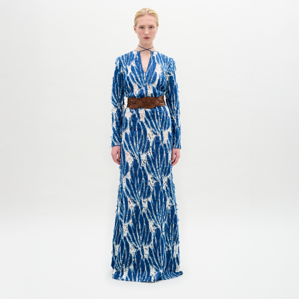 A person stands against a plain background wearing the Ebba Dress, a long blue and white patterned dress with a brown belt, high neckline, and subtle criss-cross detailing that enhances its elegant column silhouette.