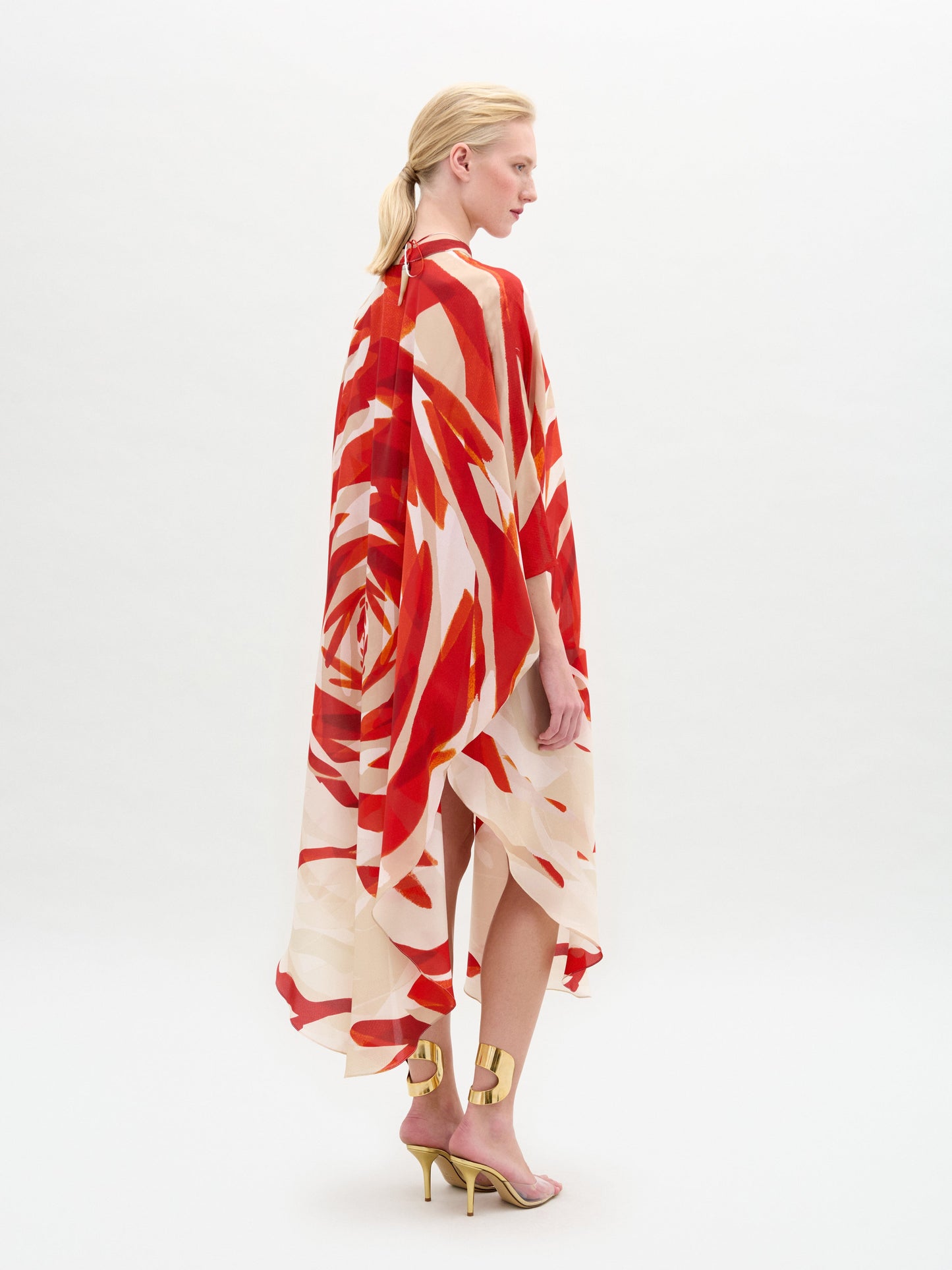 A woman stands against a white background wearing the Elea Tunic, a loose, knee-length dress made from Italian fabric with a red and cream spiral pattern. She holds a beige clutch and pairs the look with gold high-heeled sandals.