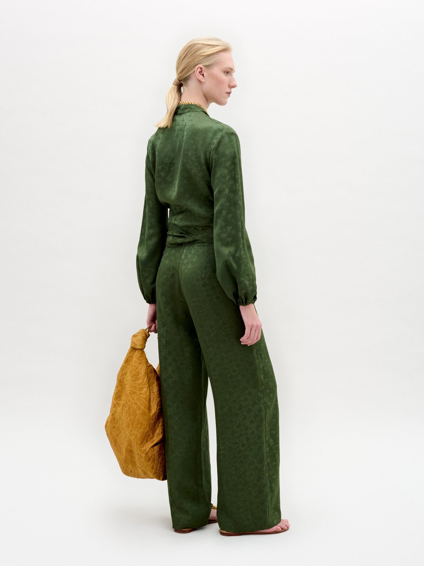 Person standing against a plain background wearing the green Elvie Blouse, a long-sleeved wrap top with wrap-around ties, matching pants, accessorized with a gold necklace and holding a gold handbag.