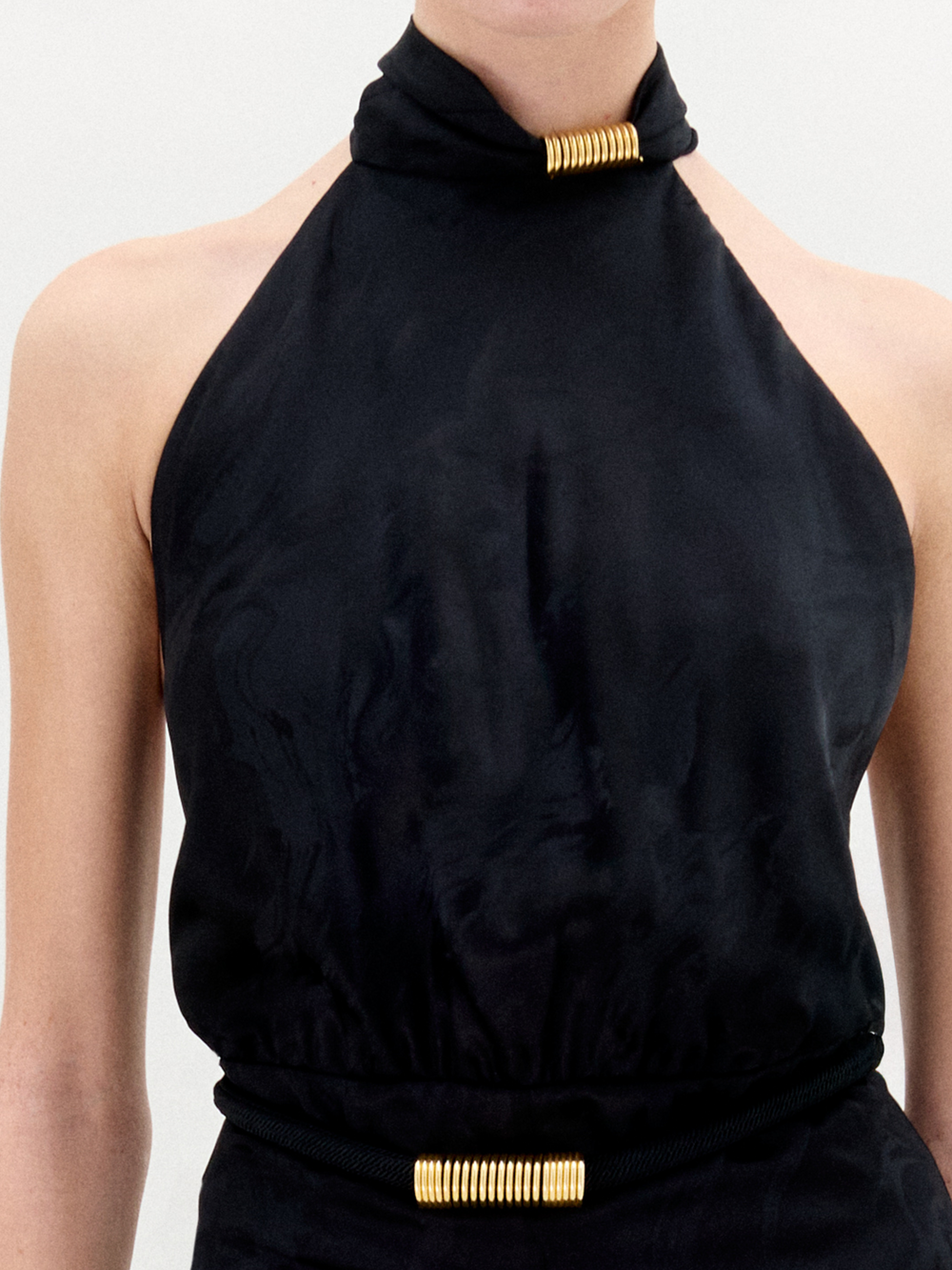 A person stands against a white background in the Enara Jumpsuit, a black halter-neck piece made from luxurious Italian fabric with wide-leg pants, accessorized with elegant gold bangles.