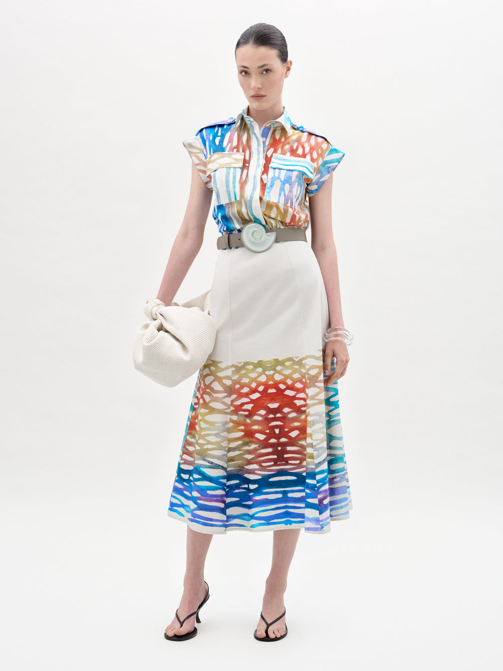 A woman wears the Erin Skirt, a colorful, abstract-patterned midi with an A-line silhouette, paired with a matching blouse, wide belt, white bag, clear bangles, and black sandals against a plain background.