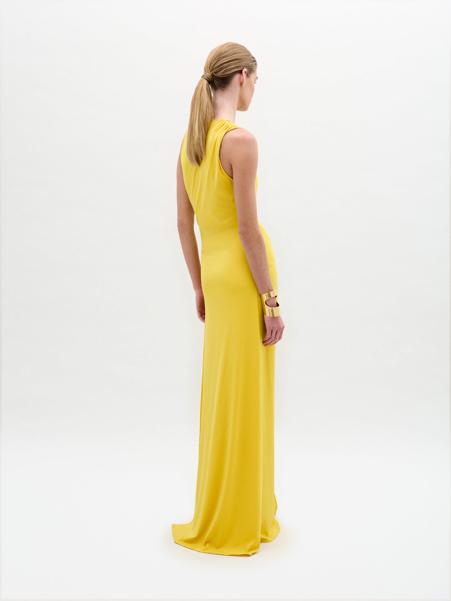 A person models the Fadila Dress, a sleeveless yellow maxi with a fitted silhouette, deep V-neckline, and striking gold hip embellishment, standing against a plain white background.