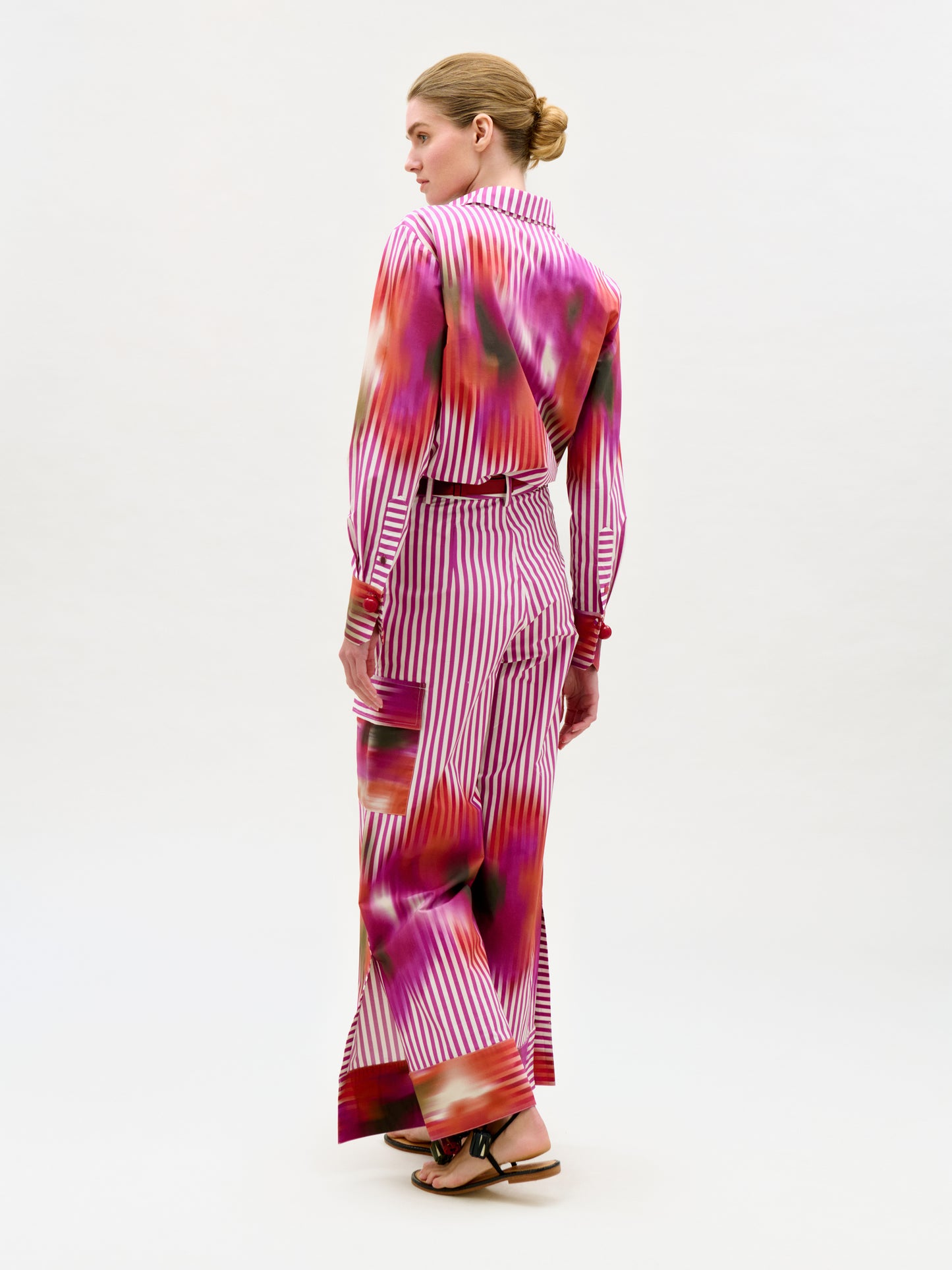 A woman faces away, wearing the Falak Pant—crafted from luxurious Italian fabric with magenta, red, and white stripes and splashes. Her hair is in a neat bun, and she wears black sandals.