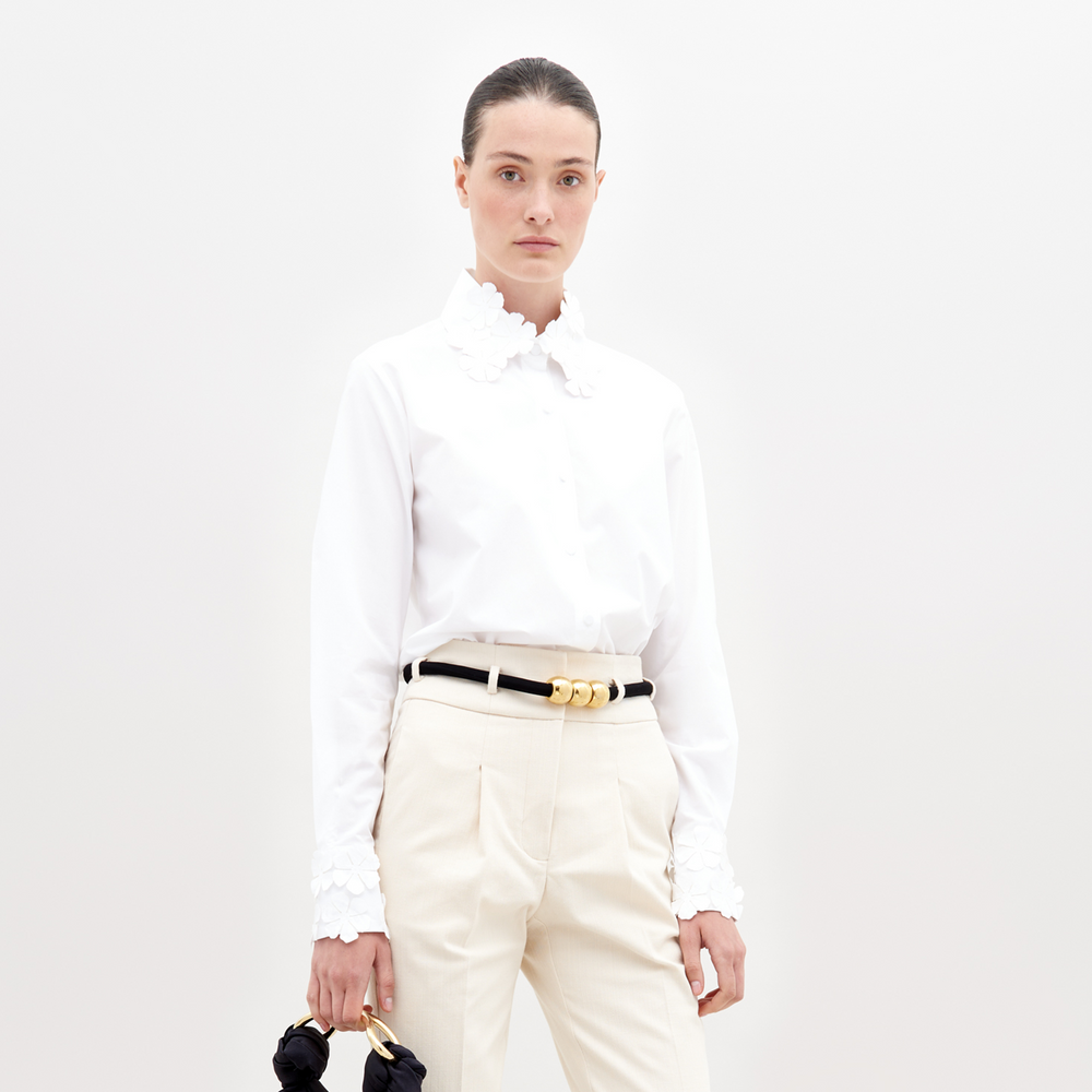 A person with dark hair tied back, wearing a Flower Blouse White with flower details on the collar and sleeves, beige pants, and a black belt with gold accents, holding a small black handbag. Pre-order now to ensure your outfit ships by November 15th, 2024.