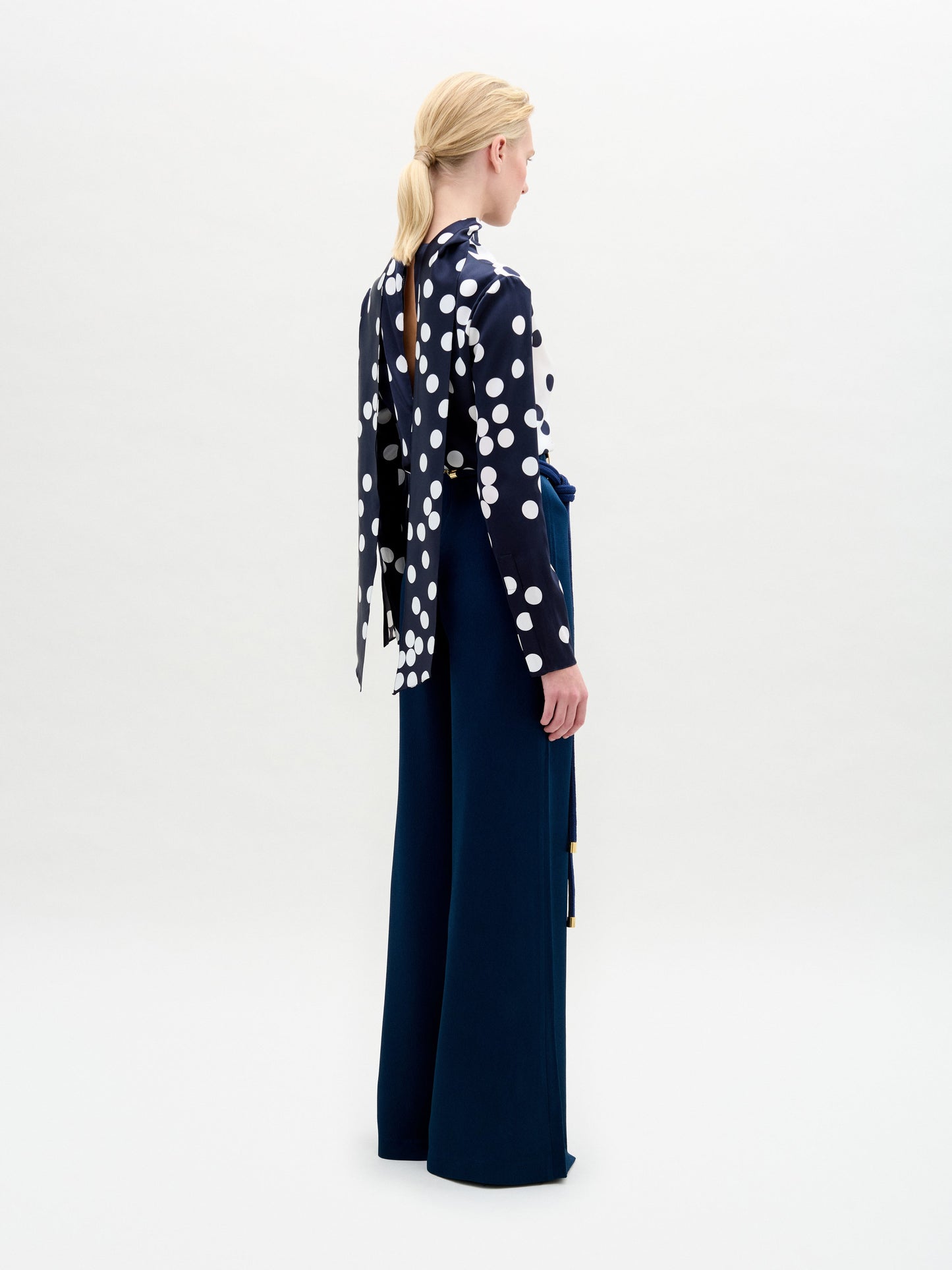 A person stands facing forward in a polka dot top with a keyhole cutout and high-waisted navy Palermo Pant featuring wide legs and a decorative gold belt, set against a plain white background.