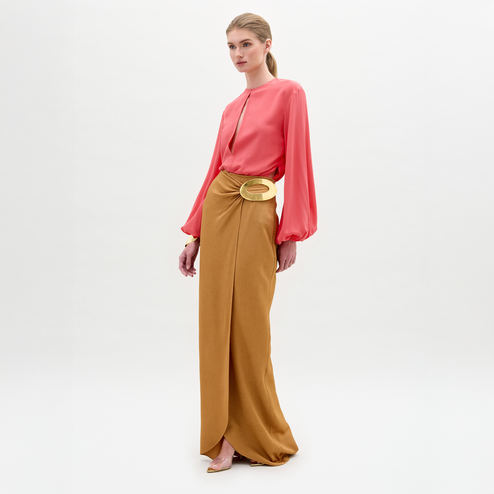 A woman stands wearing a long-sleeve coral blouse tucked into the Gadea Skirt, a high-waisted hammered satin maxi with a striking gold-tone embellishment at the waist.