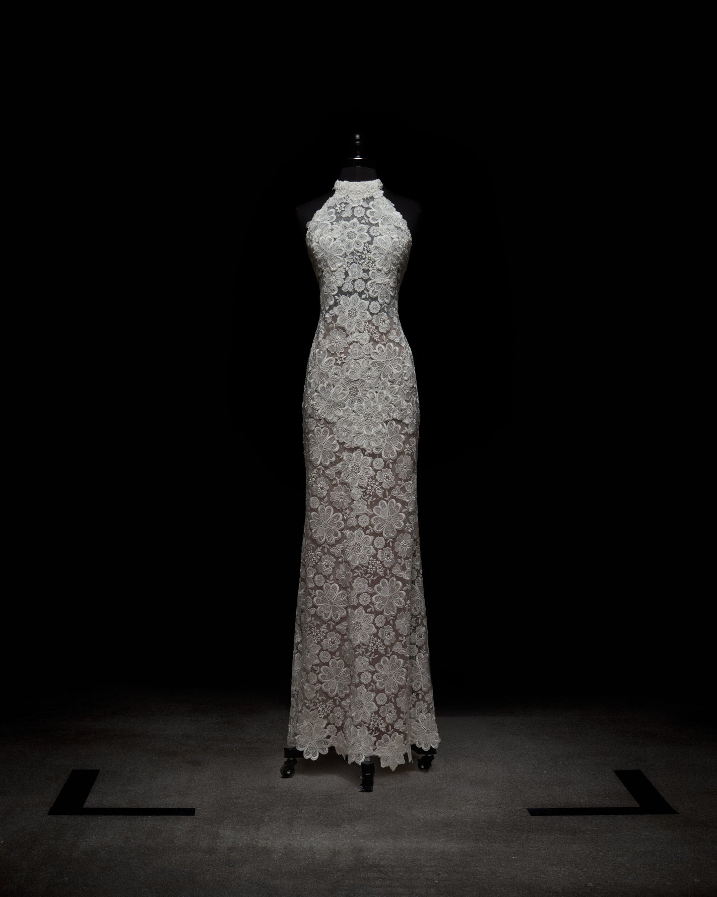 The Gardenia Dress is a floor-length, sleeveless sheer lace maxi dress with a high neckline and an intricate floral pattern, displayed on a black dress form against a dark background.