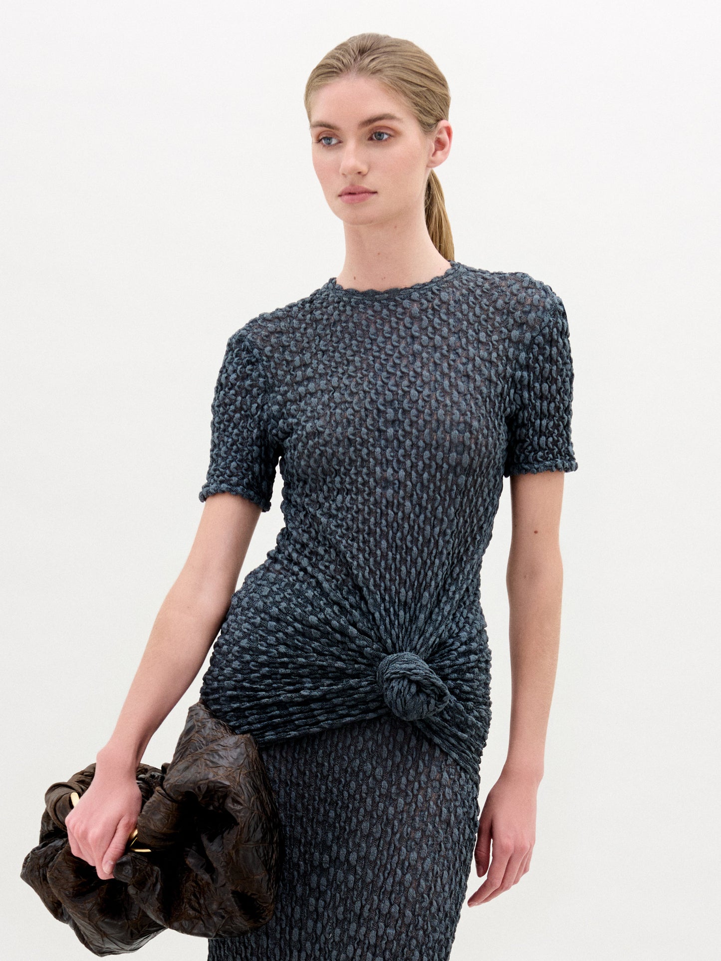 A woman with blonde hair in a low ponytail wears the Gen Tshirt, a textured, fitted, dark short-sleeve top with knot detail, and holds a large dark ruched bag against a plain white background.