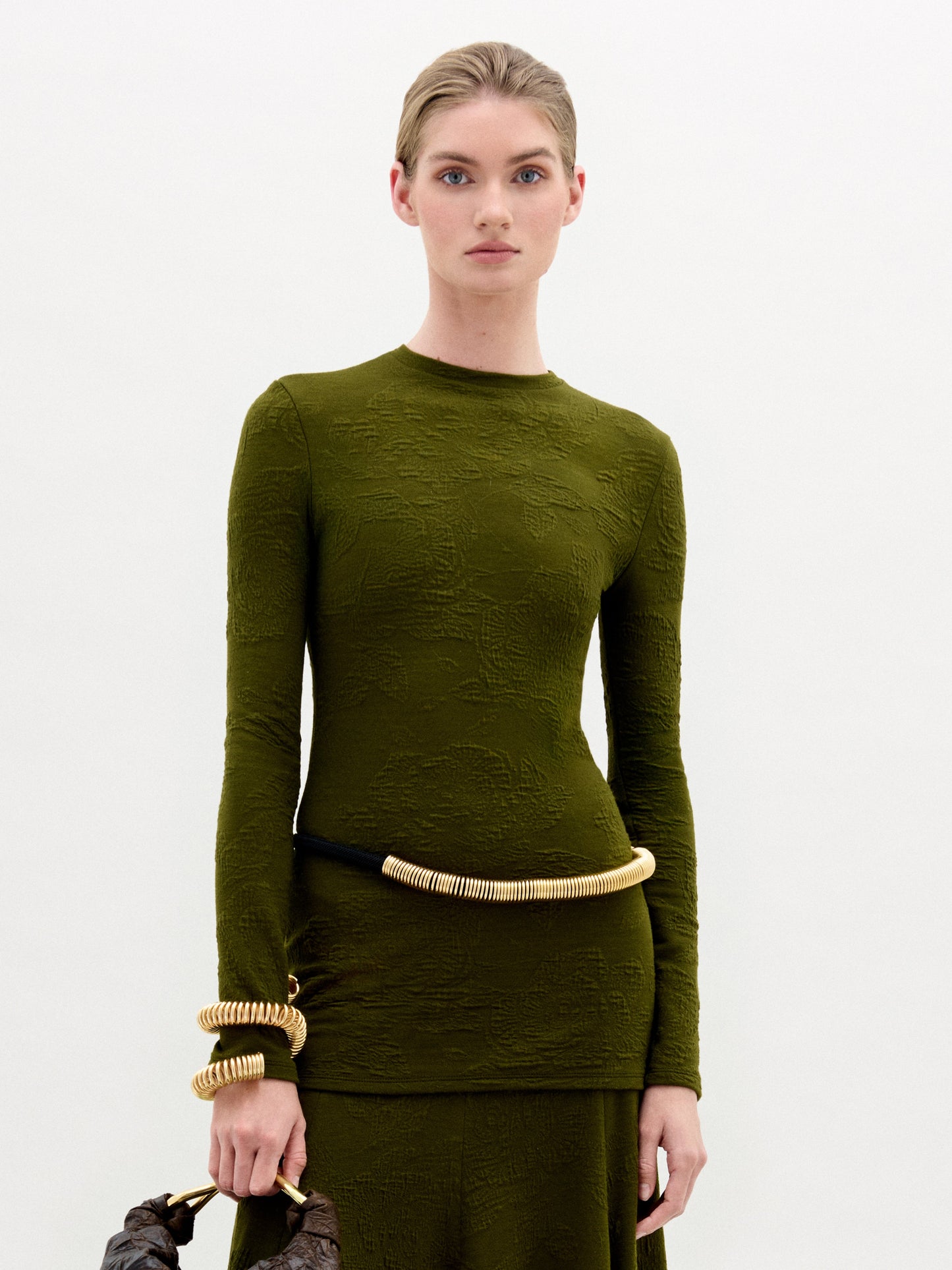 A woman stands against a plain white background wearing the Gen Tshirt, a slim fit, textured olive green long-sleeve dress with gold accessories and holding a brown bag.