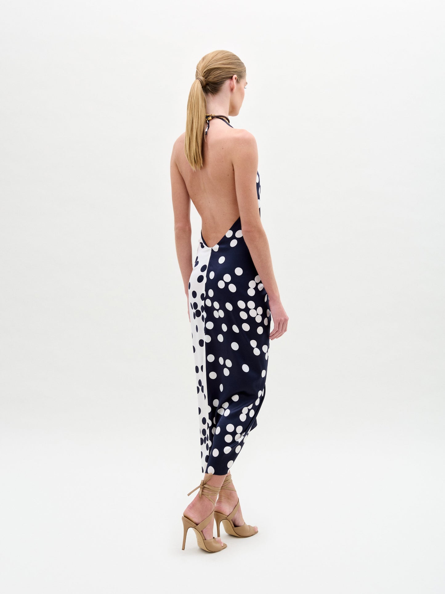 Wearing the Guadalupe Dress—a navy polka dot halter with a knotted front—paired with a colorful necklace and strappy heels, the person stands against a plain background.