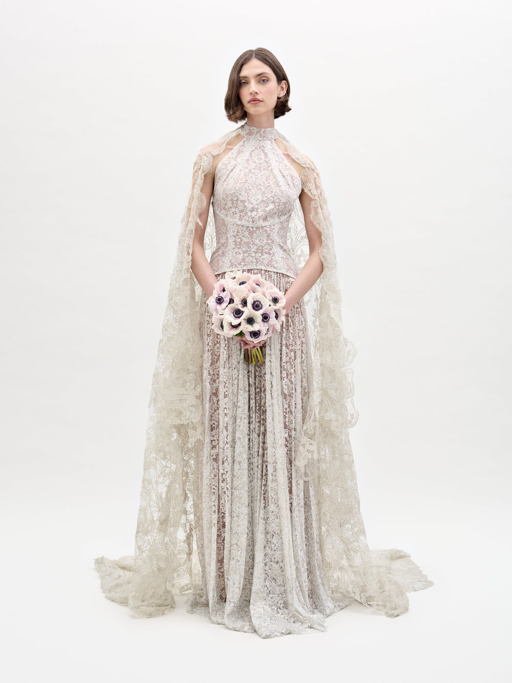 A woman wearing the Olinta Veil, a high-neck, floor-length lace gown with a scalloped hem and bridal lace cape, holds a bouquet of pale flowers against a plain white background.