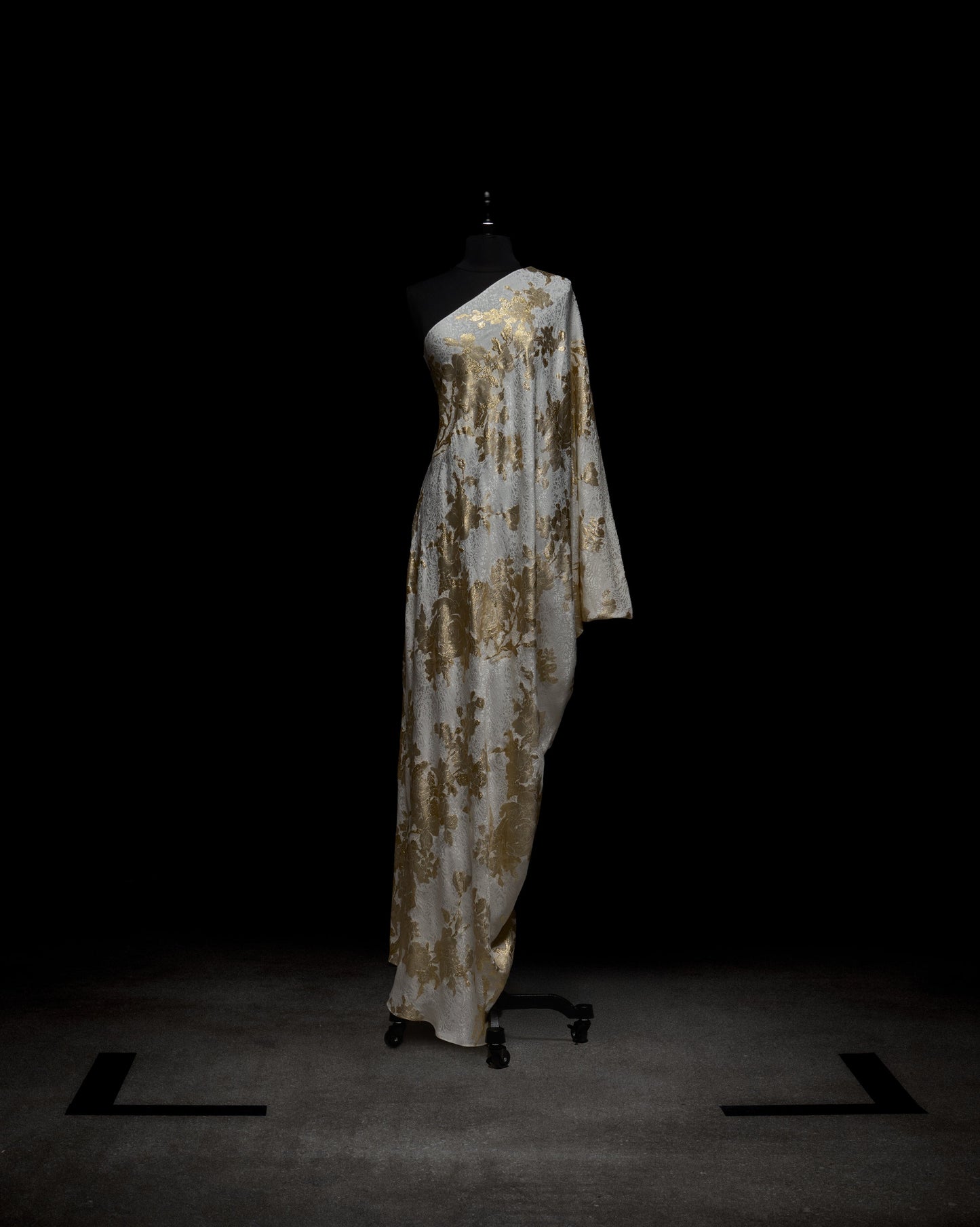 A woman dons the elegant Inu Dress, a flowing white and gold attire with a floral pattern, standing against a plain white background.