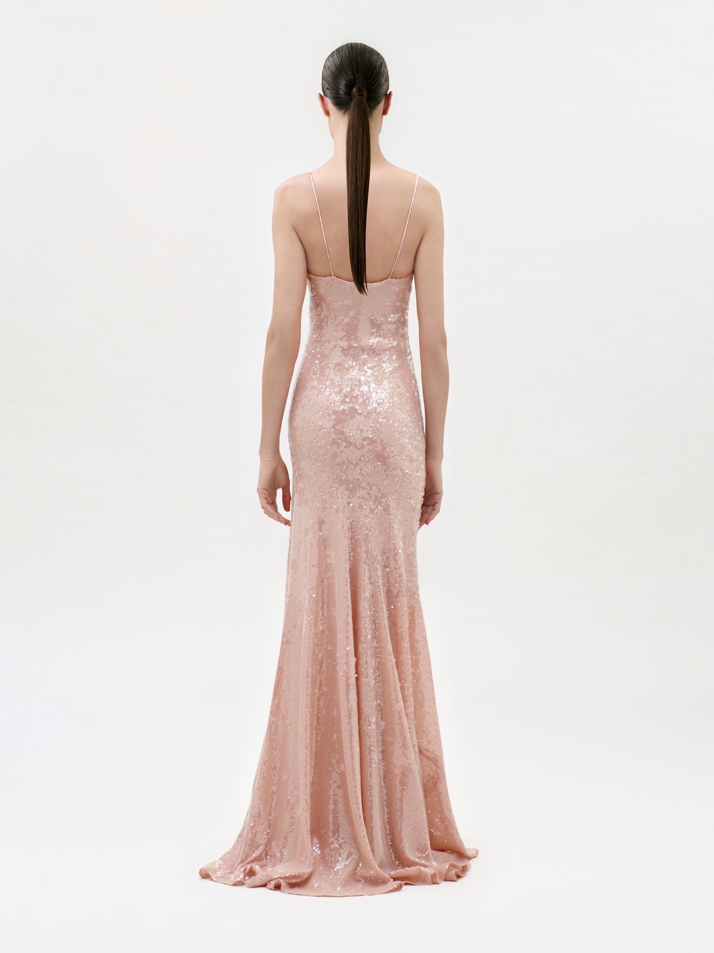 A woman with long, dark hair in a ponytail stands with her back to the camera against a plain white background, wearing the Ibona Dress—a floor-length, fitted blush pink sequin maxi dress with thin straps.