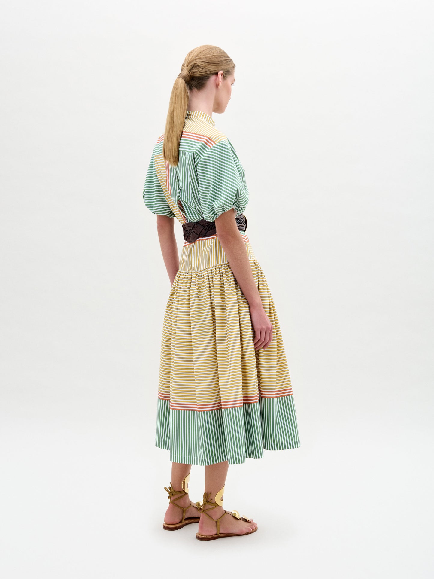 A woman stands against a plain background wearing the Jil Dress, a green and yellow striped midi with puffed sleeves, an A-line skirt, wide belt with large buckle, and gold sandals.