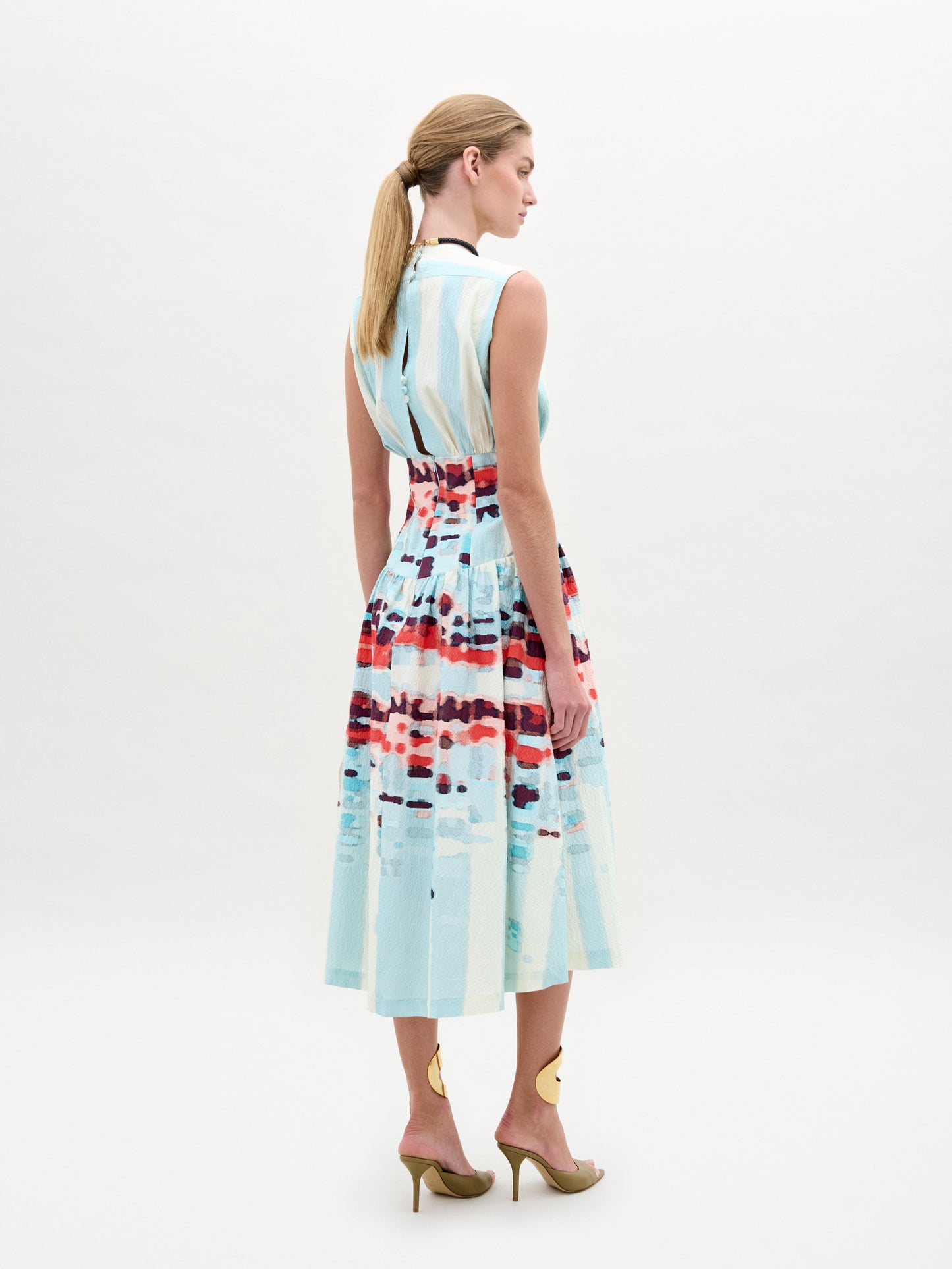 A woman poses against a plain backdrop in the Jocelyn Dress, a sleeveless V-neck midi made from fine Italian fabric with blue stripes and red patterns. She accents the look with gold sandals, a choker, and carries a beige handbag.