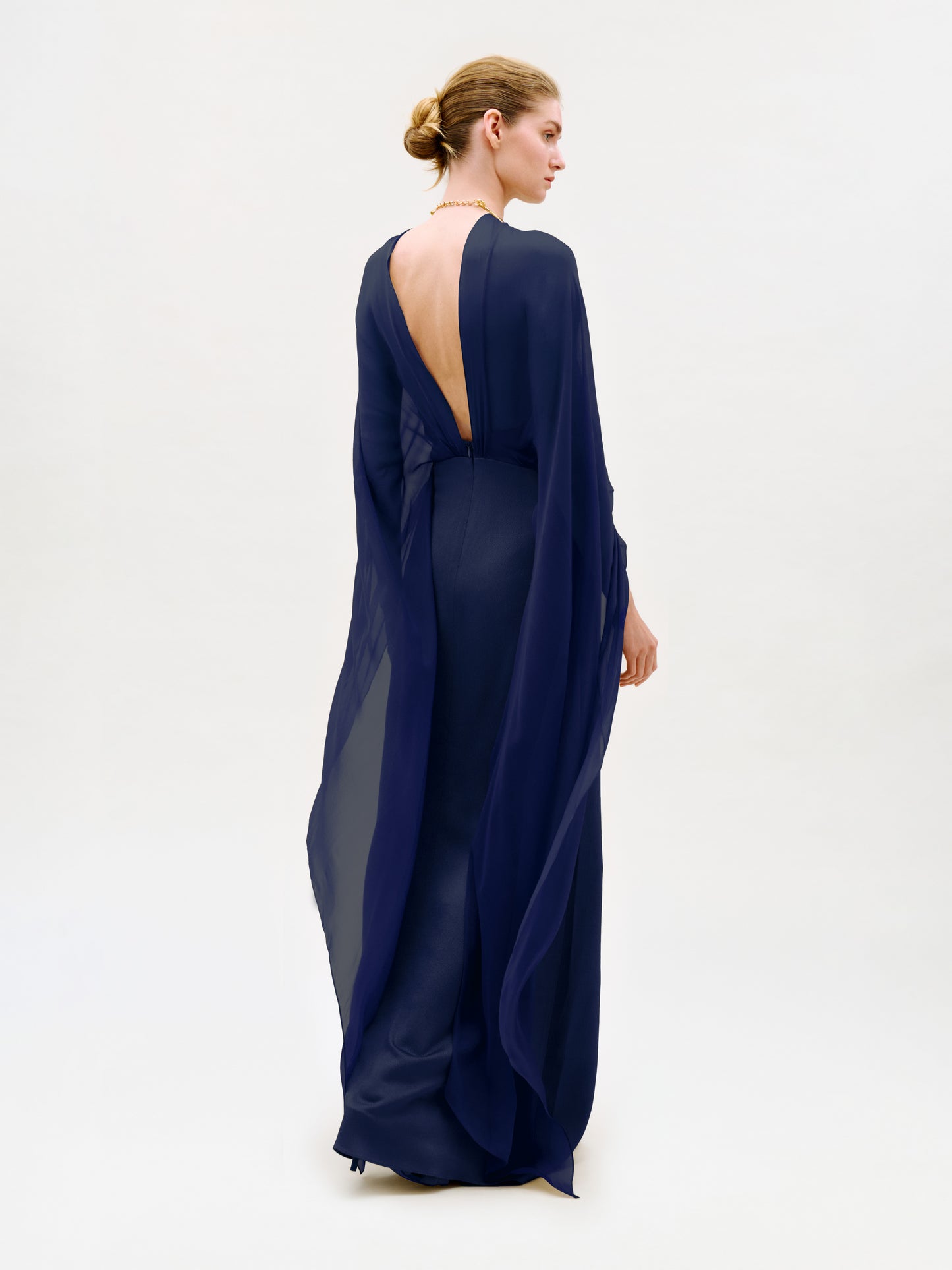 A person stands facing away, wearing the Kalina Dress—a floor-length, deep blue maxi gown crafted from luxurious Italian fabric. The dress features a sheer, flowing design with a low-cut back and column silhouette, while their hair is styled in a neat bun against a plain, light background.