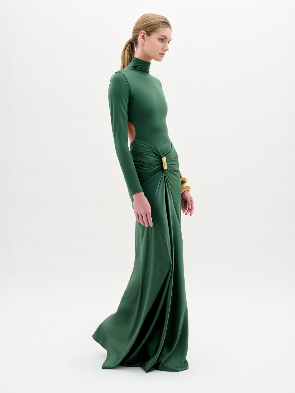 A woman wears the Lena Dress—a long green maxi with a high neck, gathered skirt, long sleeves, and side cutouts—while standing against a plain white background.