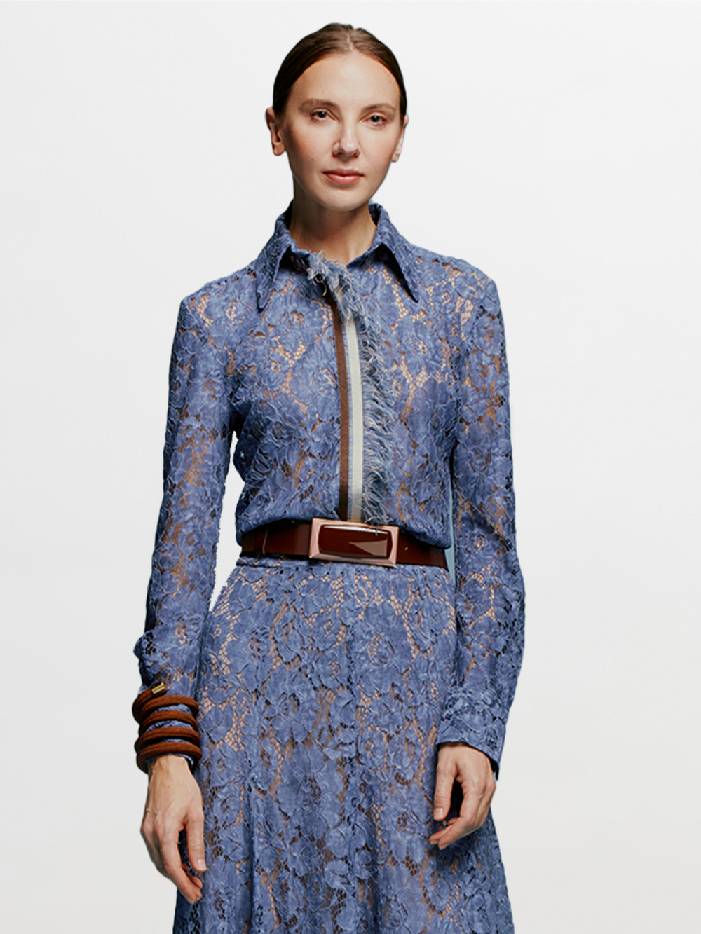 A woman stands against a plain blue background, wearing the Liv Blouse in Blue. She pairs it with a stylish brown bracelet, perfectly capturing the essence of September 2nd, 2024.
