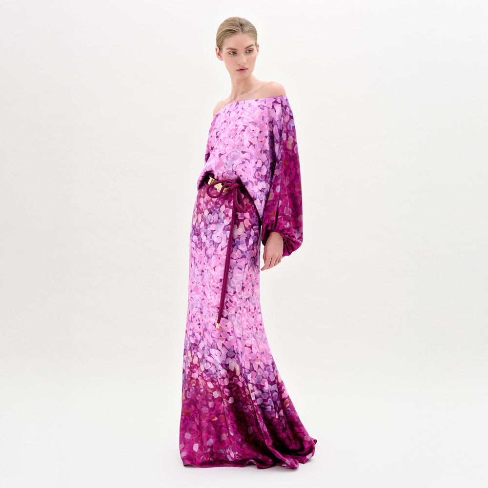 Woman models the Lorella Dress—an off-the-shoulder maxi with purple and pink florals, crafted from luxurious Italian fabric and finished with a tied belt—against a plain white background.