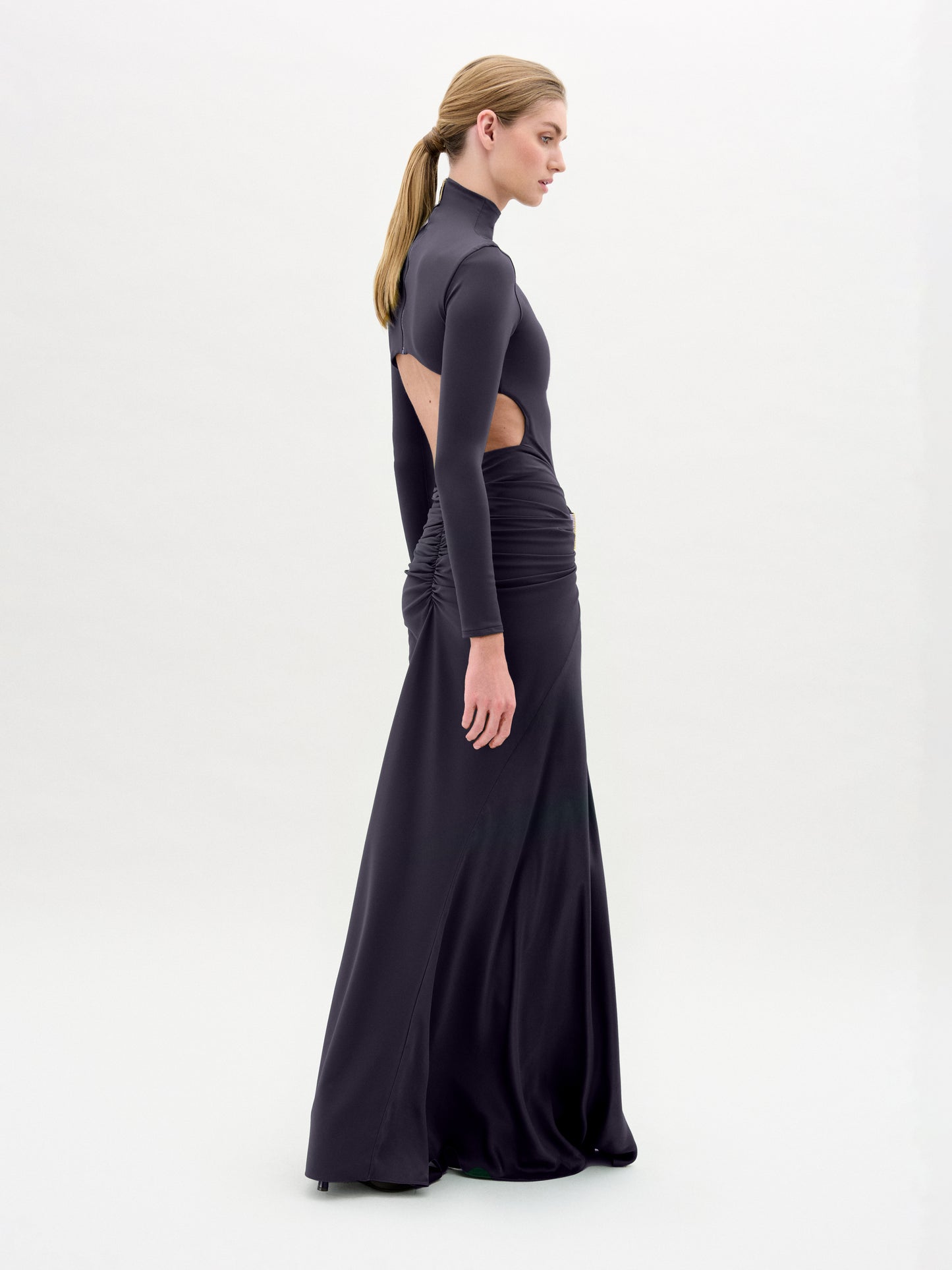 A woman wearing the Lena Dress—a long, fitted black dress with cut-out sides—stands in profile against a plain white background, her hair tied back in a low ponytail.