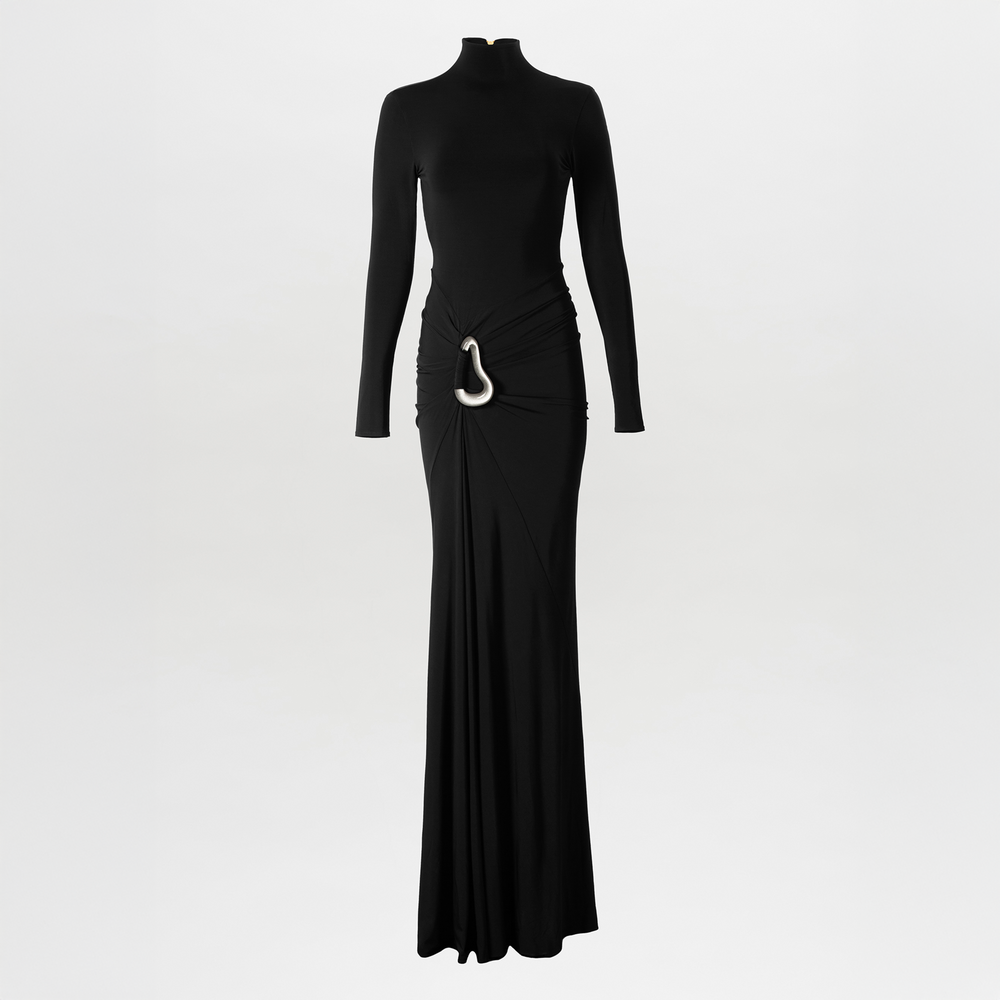 A woman poses against a plain white background wearing the Lena Dress, a long black maxi featuring a high neckline, open back, gathered waist, draped skirt, and white decorative detail at the hip.
