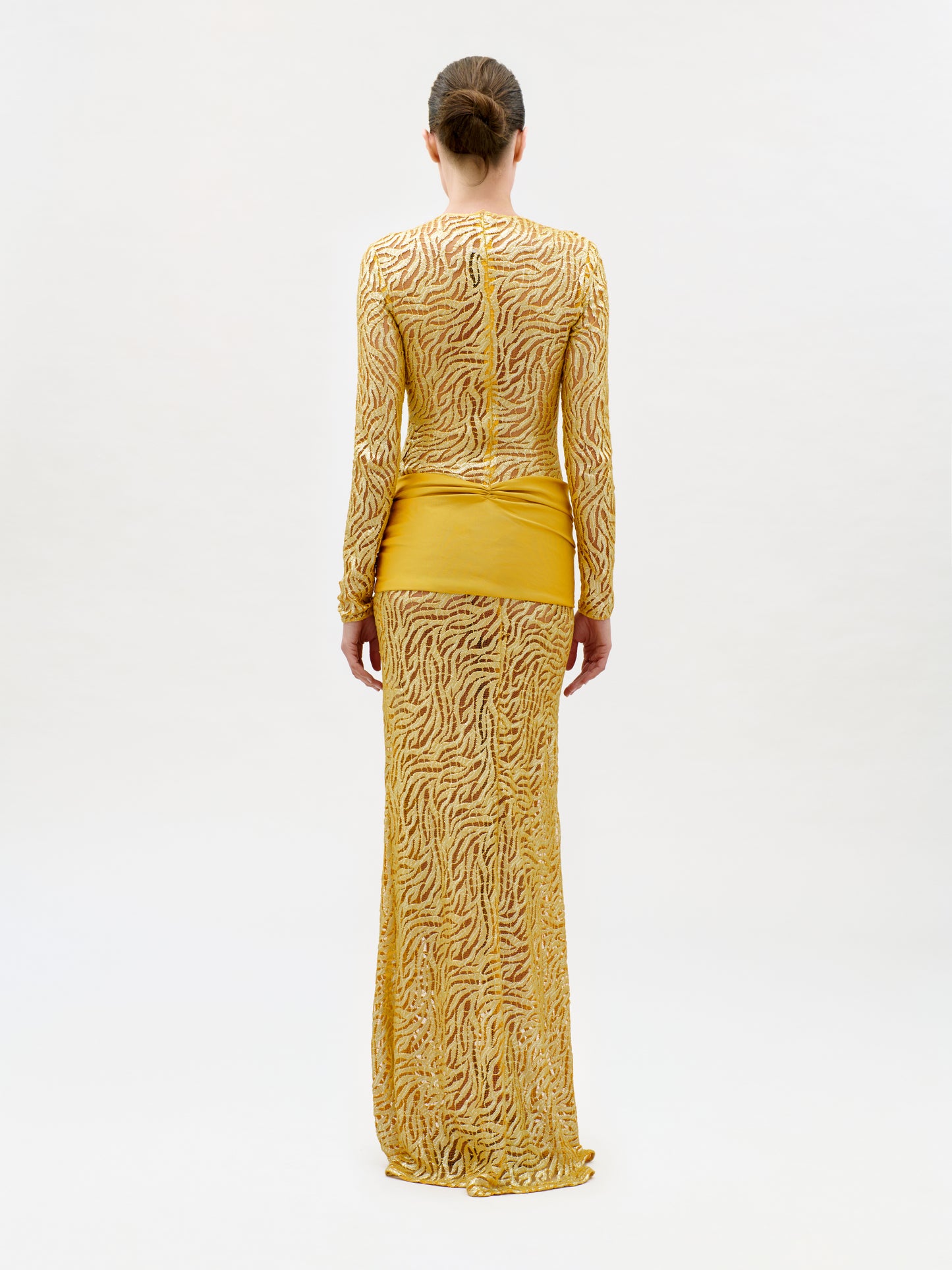 A person stands facing away, wearing the Lia Dress—a slim fit, long-sleeved, floor-length yellow gown crafted from luxurious Italian fabric featuring intricate sheer wavy patterns and a solid yellow panel around the hips. The background is plain and light-colored.