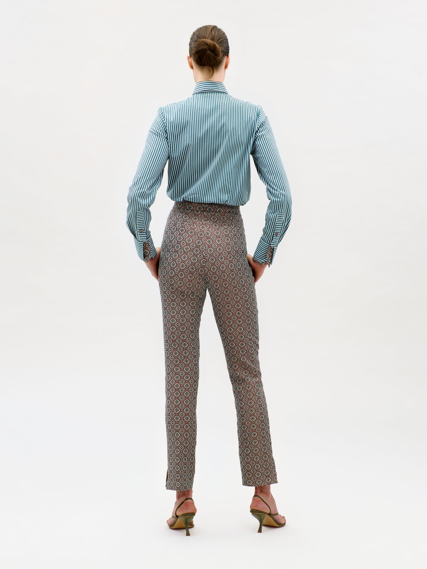 A person stands facing away against a plain white background, wearing a blue striped shirt tucked into the high-waisted Luca Pant made with premium Italian fabric, paired with heeled sandals and hair tied back.