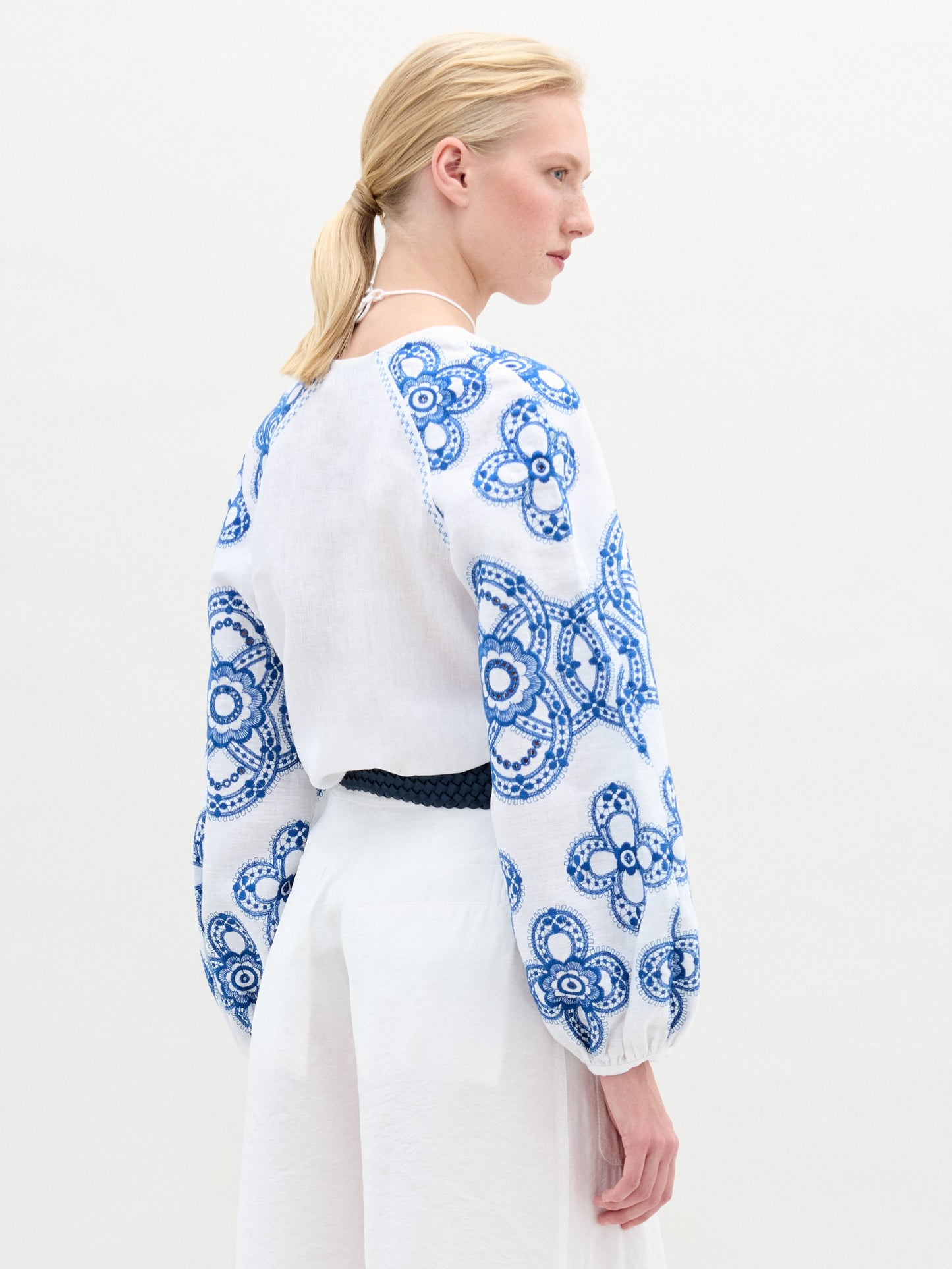 A woman stands against a plain background wearing the Molveno Blouse, an embroidered linen top with blue detailing and a V neckline, paired with a white skirt featuring a pocket and finished with a dark braided belt at her waist.
