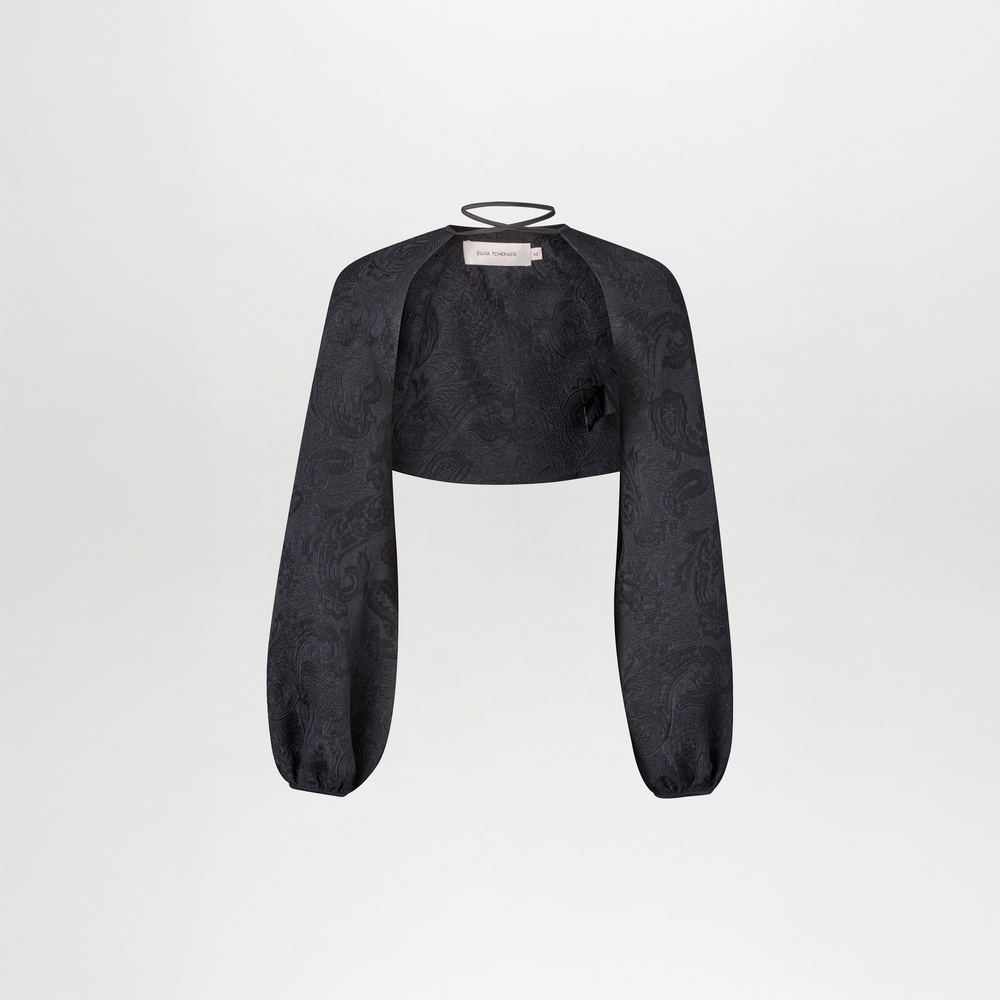 The Nana Cape is a black cropped bolero jacket featuring long, loose sleeves, a subtle jacquard paisley pattern, and a thin neckline strap, shown against a plain white background.
