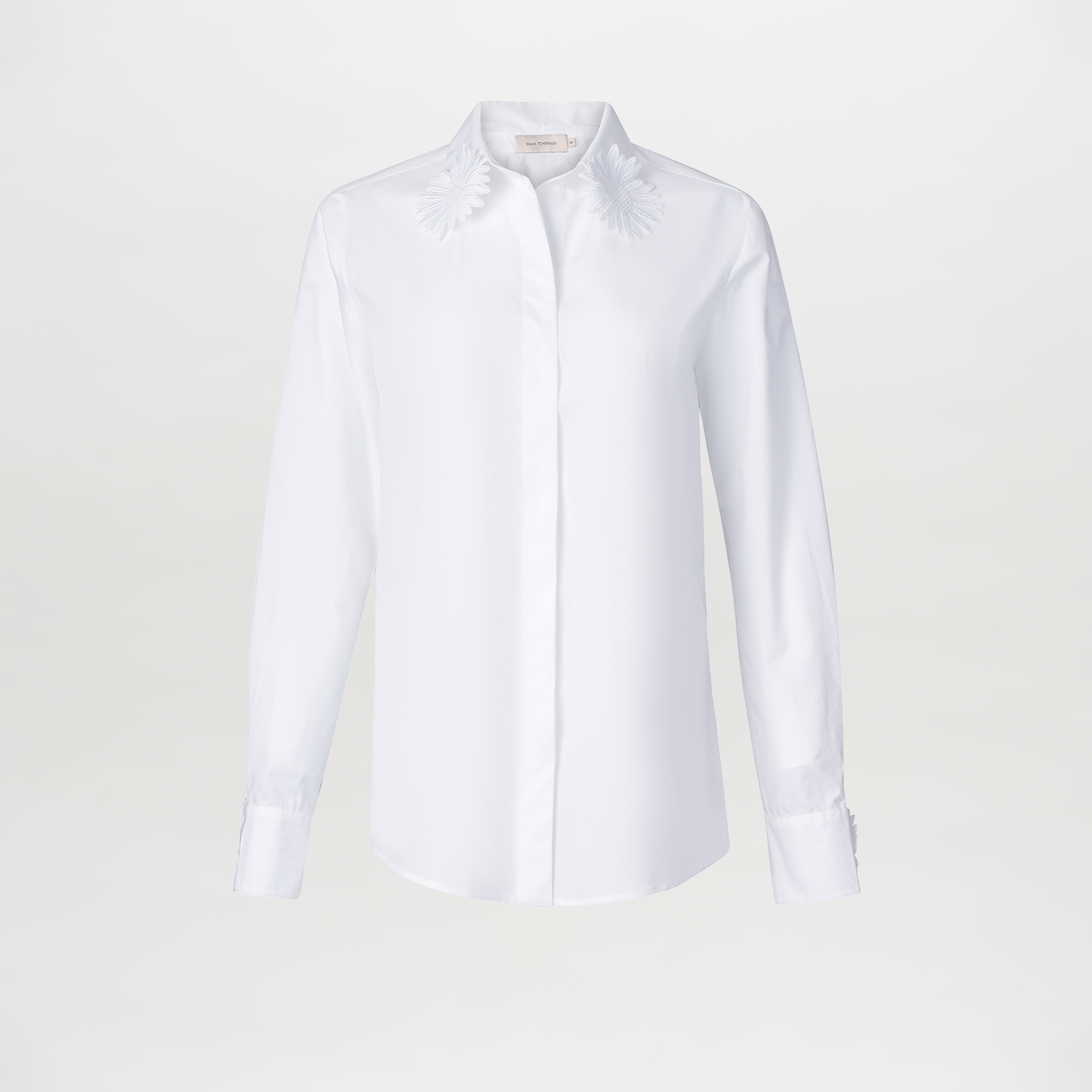 The Naroa Blouse is a white cotton top with long sleeves, a classic collar, and delicate embroidered floral details on the collar tips. Made in Colombia, it’s shown against a plain white background.