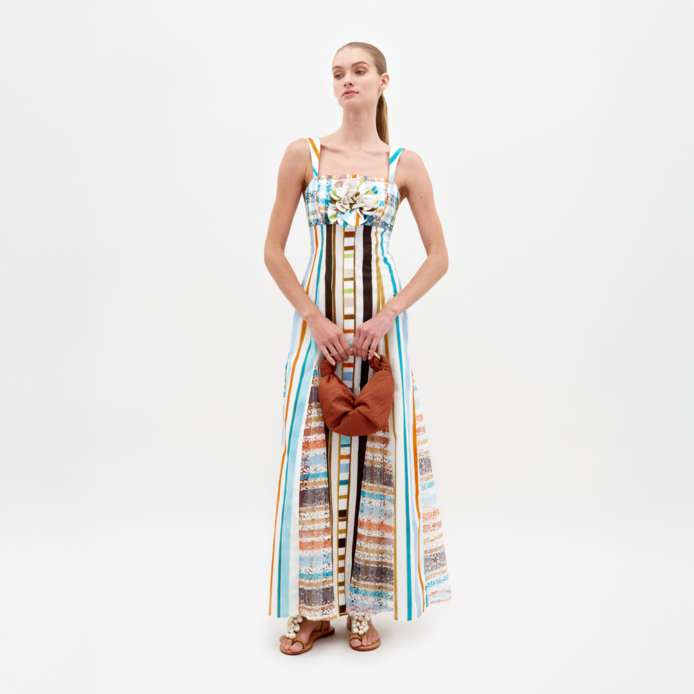 A woman stands wearing the Ofra Dress Multi Color Stripes, a sleeveless outfit featuring vibrant stripes and floral details, while holding an orange bag. The background is plain white. Please note this item is available for pre-order and will ship by February 15th, 2025.