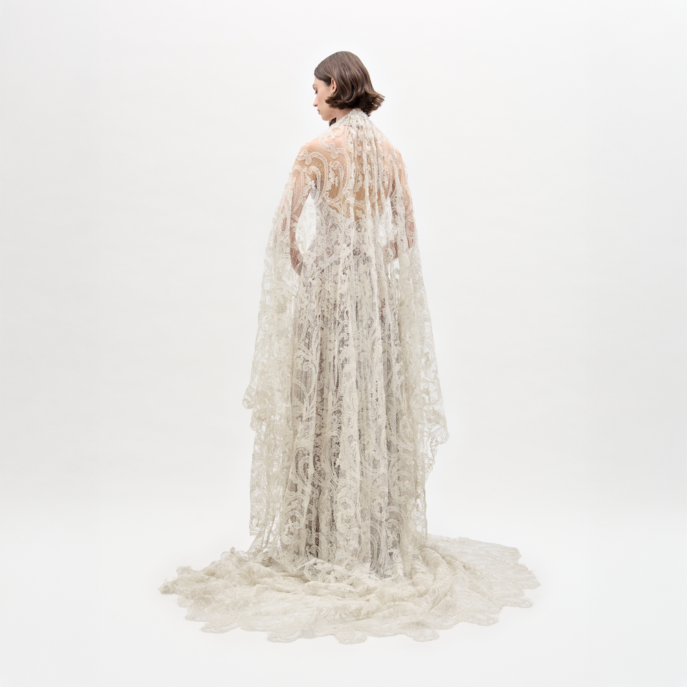 A woman wearing the Olinta Veil, a high-neck, floor-length lace gown with a scalloped hem and bridal lace cape, holds a bouquet of pale flowers against a plain white background.