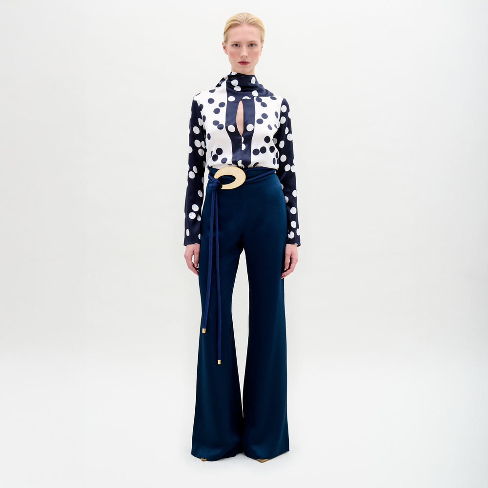 A person stands facing forward in a polka dot top with a keyhole cutout and high-waisted navy Palermo Pant featuring wide legs and a decorative gold belt, set against a plain white background.