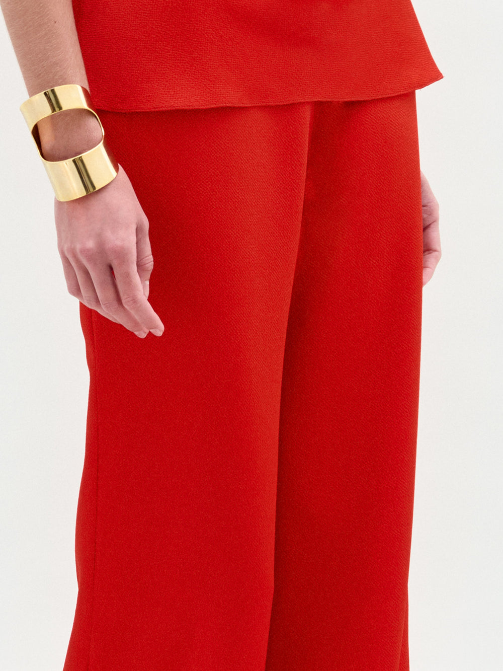 A woman poses against a white background in the sleeveless red halter top and high-waisted, wide-leg Palermo Pant in hammered satin, accessorized with gold bracelets.