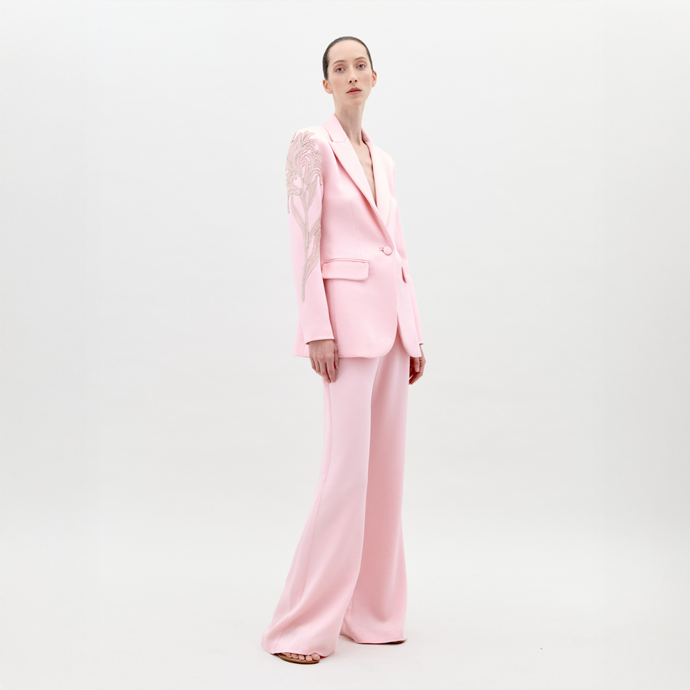 A person stands wearing the Palermo Pant Pink pantsuit, featuring floral detailing on the left shoulder and sleeve, against a plain white background. Available for pre-order now, this elegant outfit will have an exclusive ship-by date of February 1st, 2025.