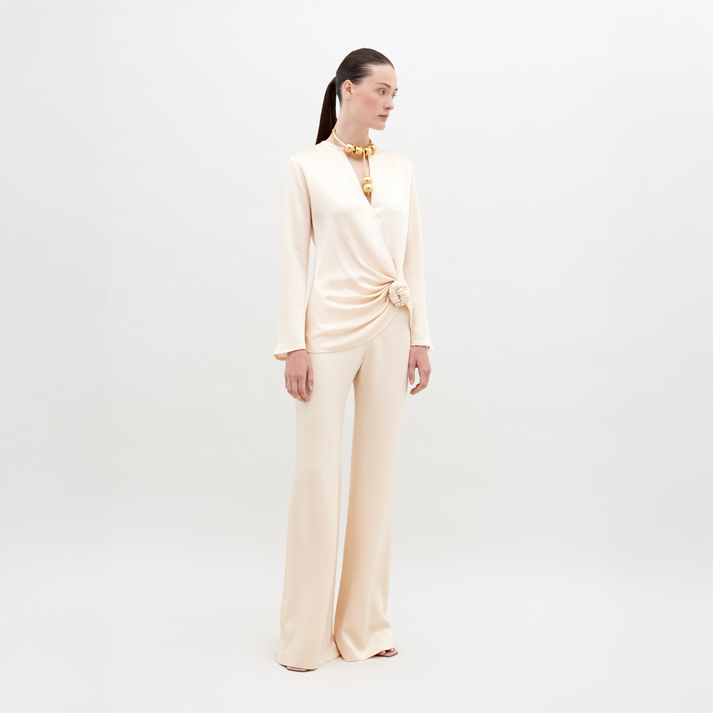 A woman stands against a plain background wearing an off-white, long-sleeve top with a knotted detail and matching Palermo Pant Vanilla. She has a high ponytail and is accessorized with a chunky necklace. Mark your calendars for November 15th, 2024—the ship by date for pre-orders of this chic ensemble.
